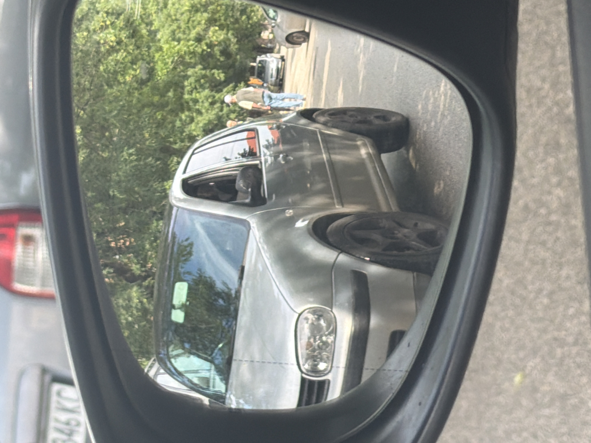 The image is a photograph taken from inside a car, showing the reflection in its passenger-side side mirror. The entire image is rotated 90 degrees clockwise.
In the reflection, a silver or light grey four-door hatchback, consistent with a Volkswagen Golf Mk4 model, is prominently featured. This car is stationary on what appears to be an asphalt road or parking area. It is equipped with black aftermarket wheels. Two men are visible standing near the reflected car; one is clearly seen wearing a light-colored cap, a vest over a light shirt, and blue jeans, while the other is partially obscured behind him. They appear to be casually standing or conversing. Further in the background, other vehicles, including another car and a motorcycle or scooter, are partially visible. The background consists of dense green foliage and trees, suggesting an outdoor, possibly roadside or park-like setting.
The perspective captures the typical distortion of a passenger-side mirror, making objects appear closer than they are. On the left side of the frame, outside the mirror's boundary, parts of the car from which the photo was taken are visible, including a red taillight and a portion of its rear license plate. The visible text on the license plate reads "346 KC". In Bulgarian license plate conventions, "KC" designates the Kyustendil region, aligning with the provided location context of Iskar, Bulgaria.
The scene takes place during daytime, indicated by the clear, natural lighting and shadows. The weather appears bright and sunny, with no signs of rain. No specific event or heightened emotions are evident; the overall impression is a mundane, everyday street or roadside scene.