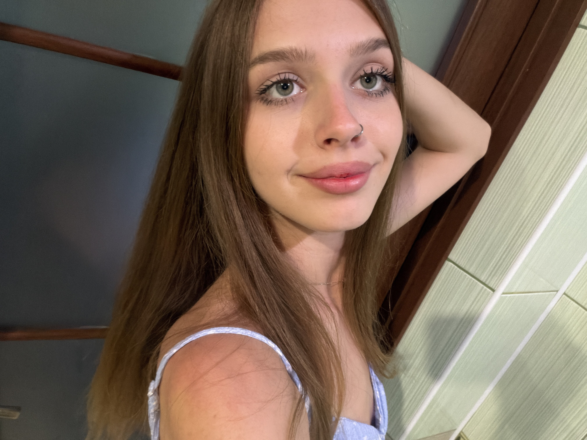 A young woman with long, light brown hair and light green eyes poses for a selfie. She is wearing a light blue, floral-patterned top. Her left arm is bent, with her hand resting on the back of her head, and she is looking directly at the camera with a slight smile. She has a small silver hoop piercing in her left nostril. The background is blurred, but appears to be an indoor setting with dark wood paneling and a tiled wall in shades of green. The lighting suggests it is daytime, but there are no external cues to determine the specific time or weather. There is no visible text in the image.