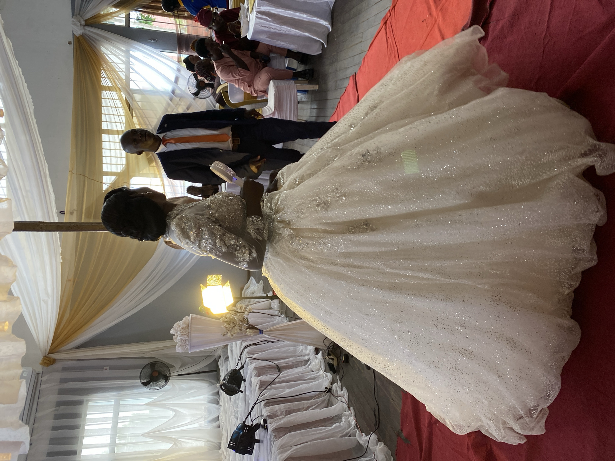 A woman in a voluminous, sparkling white wedding gown stands on a red carpet, facing away from the camera. To her right, a man in a dark suit and an orange tie looks towards her. The setting appears to be an indoor event space in Bukuru, Nigeria, decorated with draped white and gold fabric. In the background, guests are seated at tables, and a fan is visible. The lighting suggests it is daytime or a well-lit indoor event.