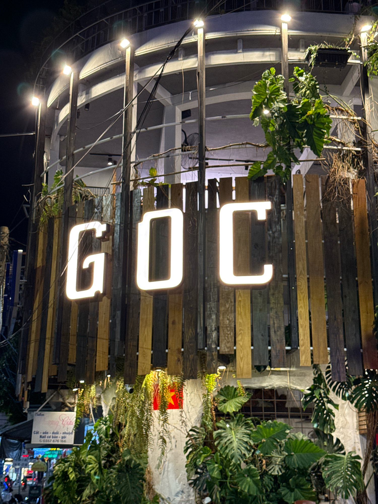 The image shows a close-up, eye-level shot of a building at night, illuminated by overhead lights and the illuminated letters "GOC" on a wooden facade. The letters are large, white, and appear to be neon or LED. The facade itself is constructed from vertical wooden planks of varying colors, from light brown to dark gray. Lush green foliage, including large-leafed plants and hanging vines, is prominently featured, partially obscuring the wooden planks and the building structure behind. Some of these plants appear to be artificial.
In the upper section of the image, the building's structure is visible, featuring a curved, multi-story design with railings and supports. Wires and cables are strung across the upper portion. The background is dark, indicating it is nighttime.
In the lower left quadrant of the image, a white sign with Vietnamese text is partially visible, along with some indistinct signs with colorful lights in the far distance, suggesting a street scene with other businesses. The overall impression is of an establishment with a natural, tropical aesthetic, likely a restaurant, cafe, or shop.
Based on visual cues such as the signage and the style of the establishment, this scene is likely located in the city of Nha Trang, Vietnam. The presence of Vietnamese text on the sign and the architectural style are consistent with this location.