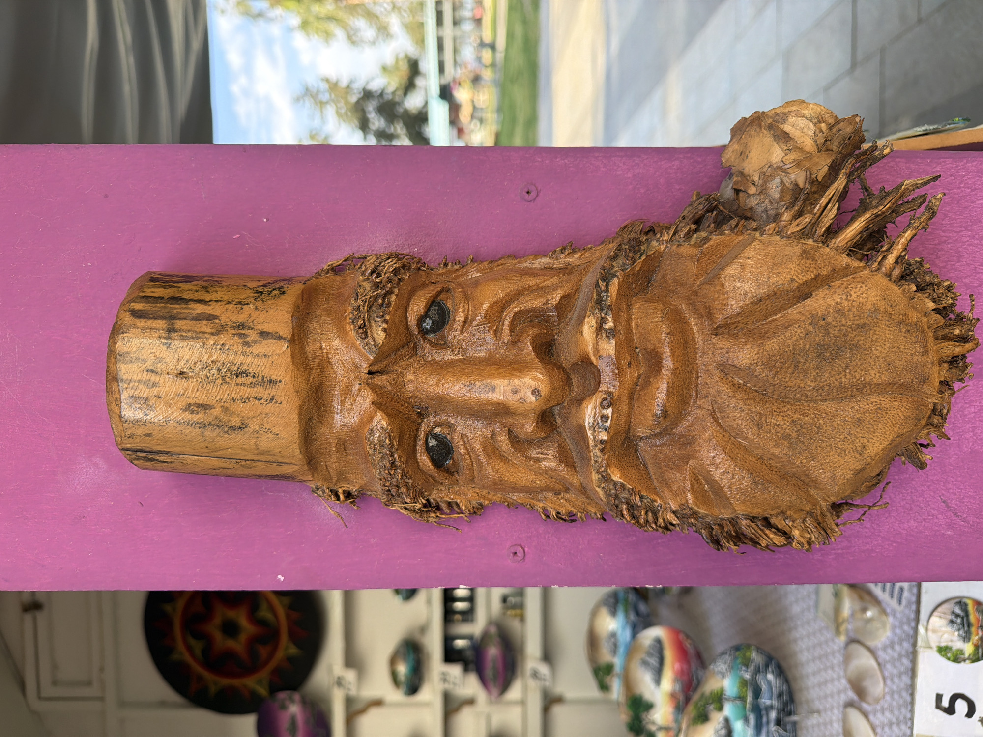 A horizontal image shows a wooden carving of a bearded man with a hat, mounted on a purple shelf or display. The carving is detailed, with defined facial features and textured beard and hair made from the same wood. The hat is cylindrical. The shelf is a vibrant purple. Below the shelf, out of focus, are various decorative items, including colorful plates or small paintings, and a rack system for displaying them. The location is indicated by the presence of items likely for sale in a tourist area, and given the prompt's details, it is assumed to be in Kemer, Turkey. The scene depicts a display of local crafts.