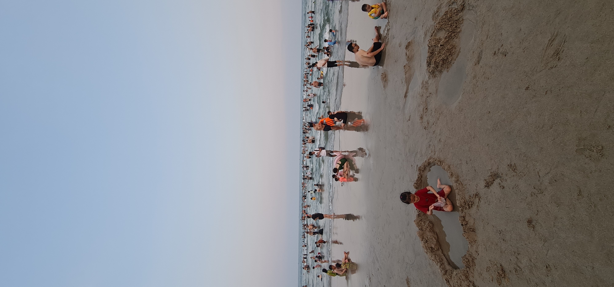 The image depicts a crowded beach in Đà Nẵng, Vietnam. A vast number of people are swimming and playing in the water. On the sandy shoreline, a child wearing a red shirt is sitting in a shallow, water-filled indentation in the sand, playing with something in their hands. Further along the beach, an adult and another child are sitting near the waterline. The sky is clear and light blue. The beach appears to be a popular destination.