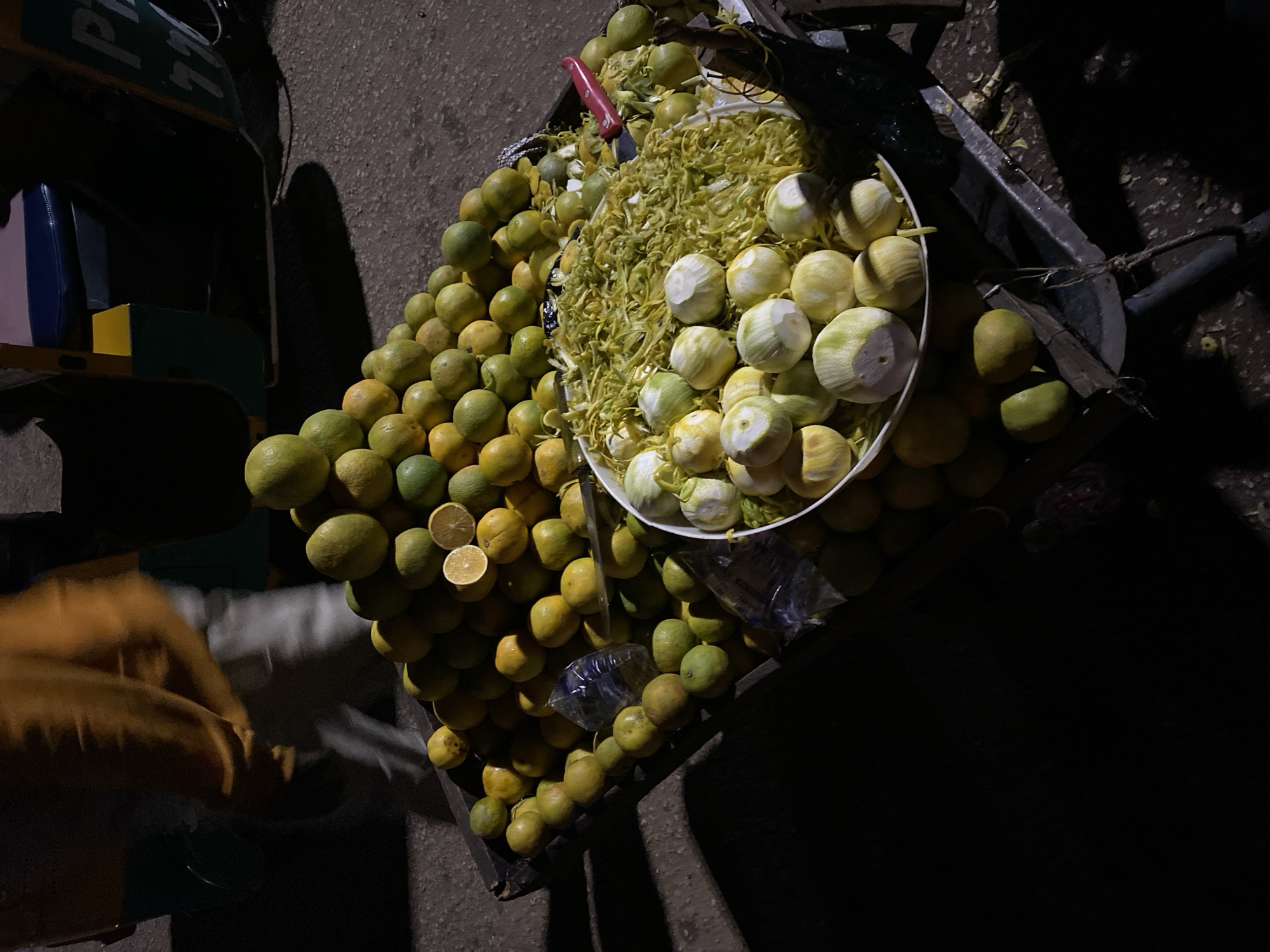 A high-angle, poorly illuminated photograph taken in Bukuru, Nigeria, shows a cart laden with citrus fruit, likely limes or lemons. The cart is positioned on a paved surface, presumably a street or sidewalk. The main focal point of the image is a mound of whole, unpeeled limes/lemons, alongside a white bowl overflowing with peeled fruit and shredded rinds. A red-handled knife is stuck into the fruit. The overall lighting is dim, casting deep shadows, and revealing sparse details of the surrounding urban environment and hinting at commercial activity related to selling citrus fruit. A blurred figure of a hand is seen on the left of the frame.