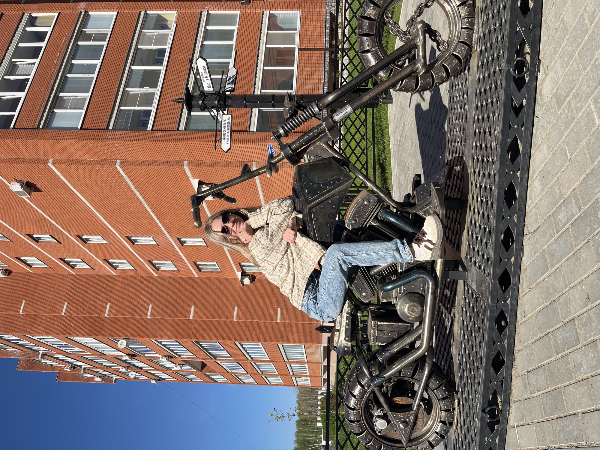 A woman sits on a metal motorcycle sculpture in Chaikovsky, Russia. The sculpture is placed on a dark gray brick surface next to a decorative metal fence. Behind the woman and sculpture is a large red brick building with numerous windows. A street sign post stands near the motorcycle with signs pointing in different directions, one of which is partially readable as "ул. Карла Маркса" (Karl Marx Street). The sky is clear and blue.