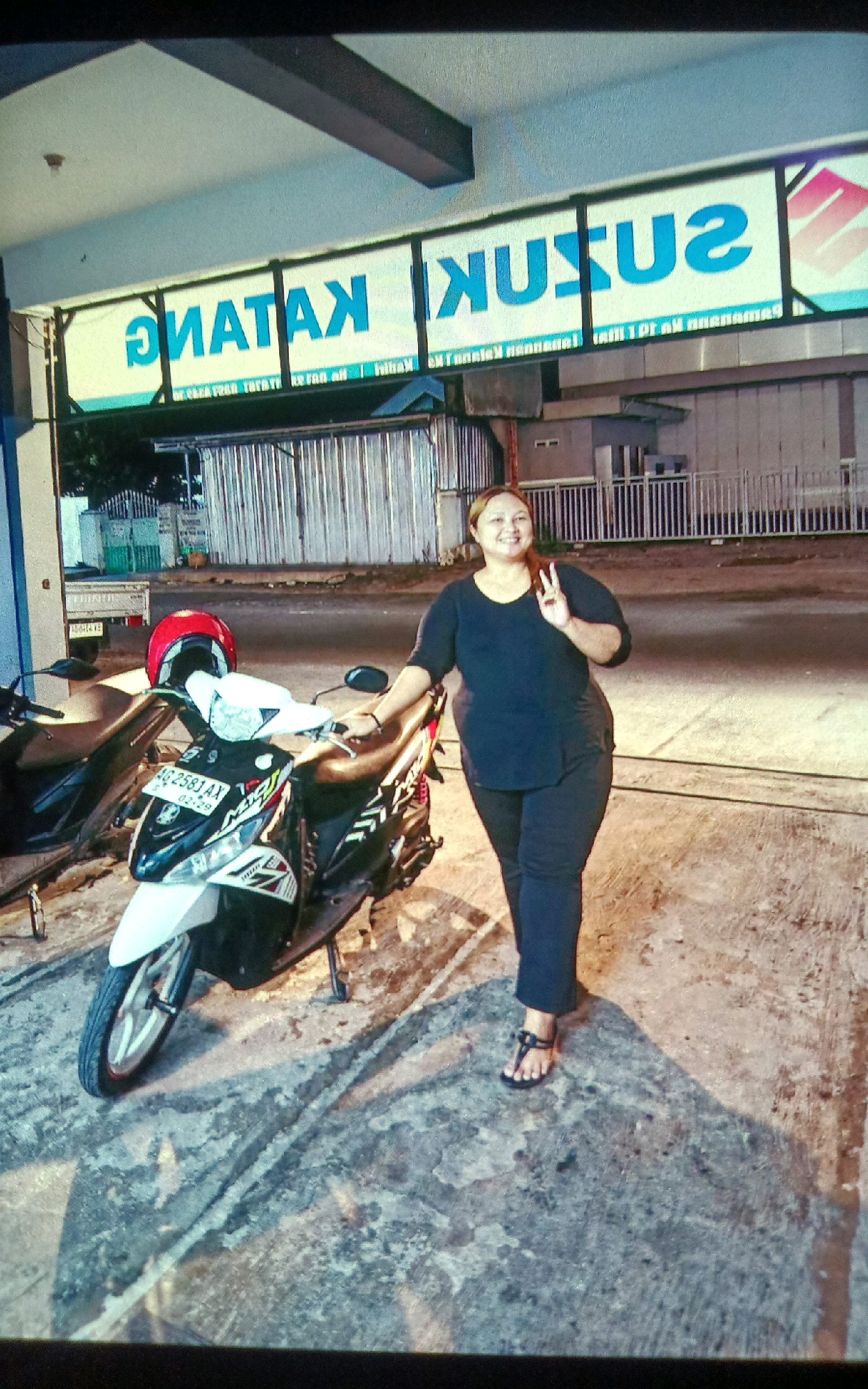 A full-body, medium shot shows a plus-size woman in a dark blue t-shirt and dark pants posing next to a white and black motorcycle parked on a concrete surface. The woman is facing slightly to the left, with her right hand raised and making a peace sign. The motorcycle is parked in front of a store with a sign that reads "SUZUKI" in blue letters, with the Suzuki logo on the right. The store appears to be located in Gampengrejo, Indonesia. The scene is at night, with artificial lighting casting shadows on the ground. A red helmet is placed on the motorcycle's handlebars.