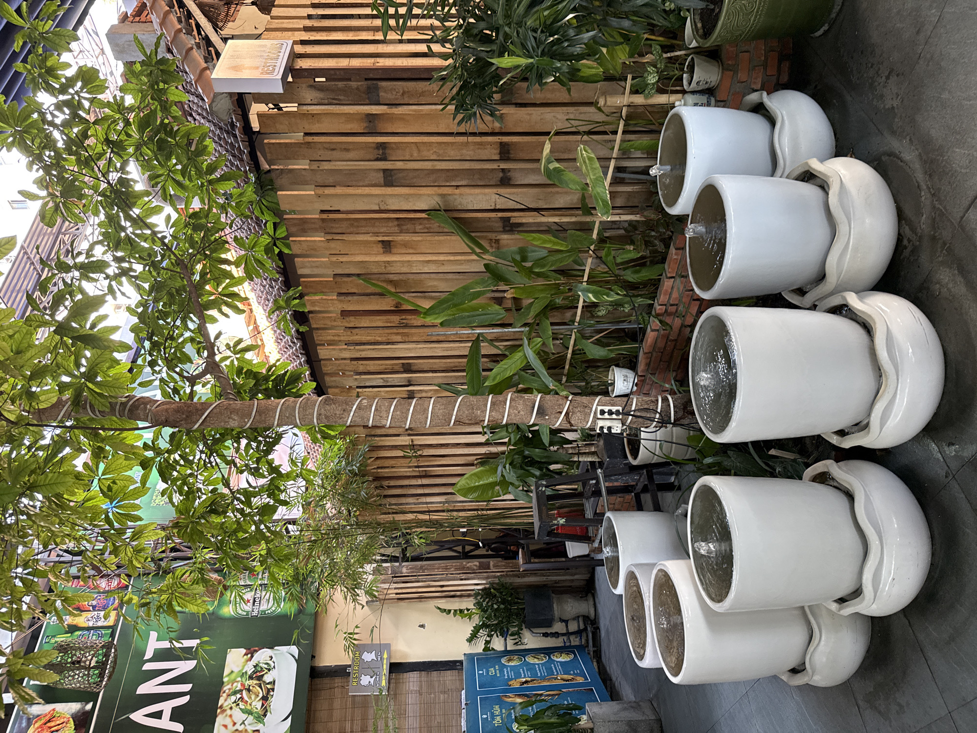 The image depicts an outdoor seating area with a rustic ambiance, likely at a restaurant in Nha Trang Ward, Vietnam, given the signage in Vietnamese. In the foreground, a line of white ceramic water fountains are arranged on a dark tiled floor. Each fountain consists of a tall, cylindrical pot with a smaller, decorative bowl at its base, and water gently cascades from the upper pot into the lower one. Behind the fountains, lush green foliage, including bamboo and broad-leafed plants, thrives. A slender tree with a trunk wrapped in white twine stands prominently to the right, its leaves casting dappled shadows.
A wooden plank fence forms the backdrop, creating a natural, textured wall. To the left, a sign in Vietnamese indicates food items. Above the fence, a glimpse of a building with a tiled roof and a sign that reads "RESTROOMS" is visible, suggesting a public establishment. The scene is bathed in natural light, indicating it was taken during the daytime. There are no people visible in the image. The overall impression is one of a tranquil, green oasis designed for outdoor dining or relaxation.