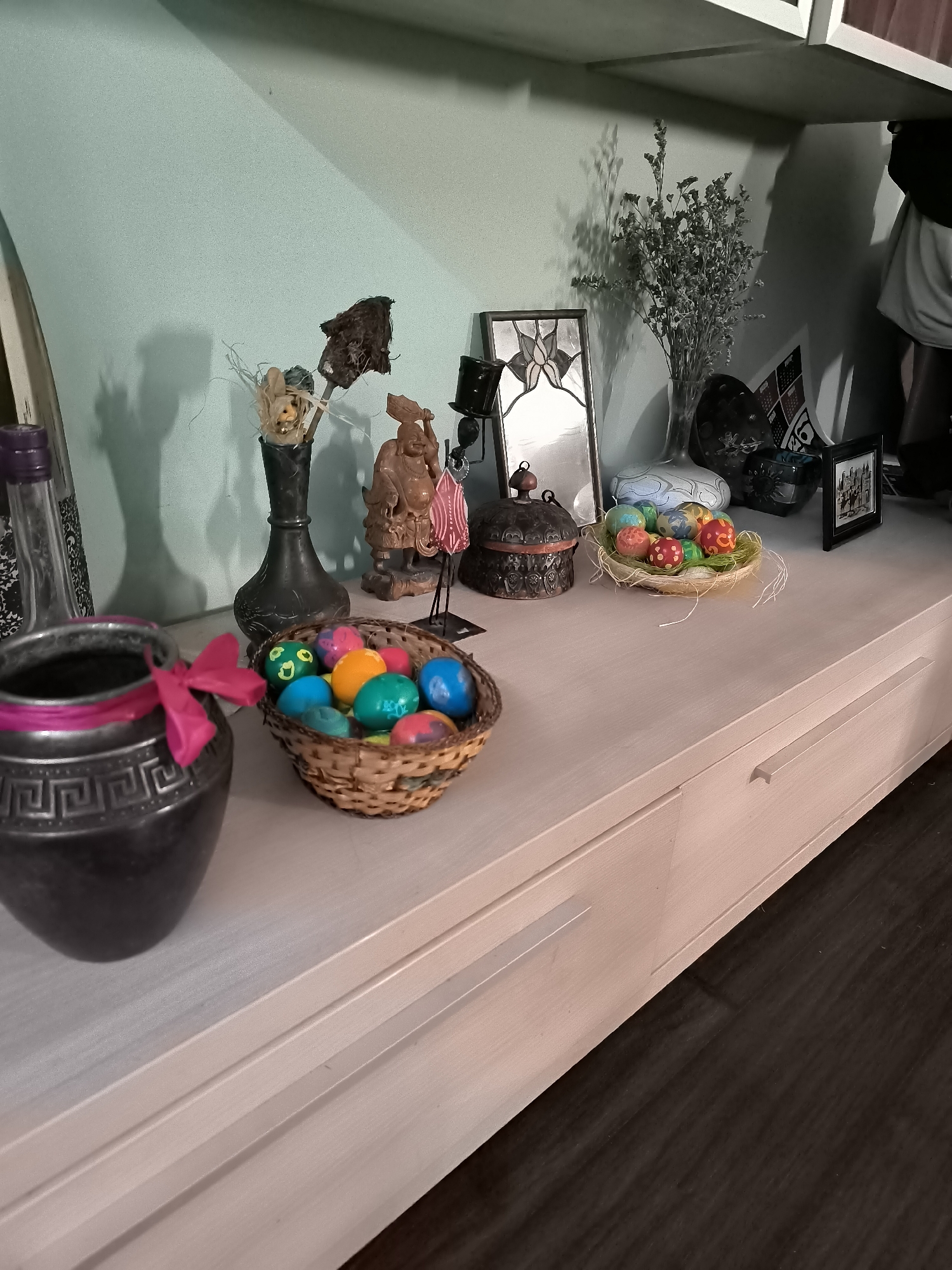A brightly lit interior setting features a collection of decorative objects arranged on a light-colored dresser or cabinet. At the left is a gray vase adorned with a pink ribbon. To its right, a small woven basket overflows with vibrantly colored Easter eggs. In the background are multiple vases and figurines of varying sizes and styles. A framed stained glass decoration sits next to a vase holding what appears to be dried flowers. A framed photograph is also present towards the right of the image. The room is tidy, with a minimal aesthetic. It is impossible to determine the location in Sofia, Bulgaria. There are no people, actions, or interactions visible in the image.