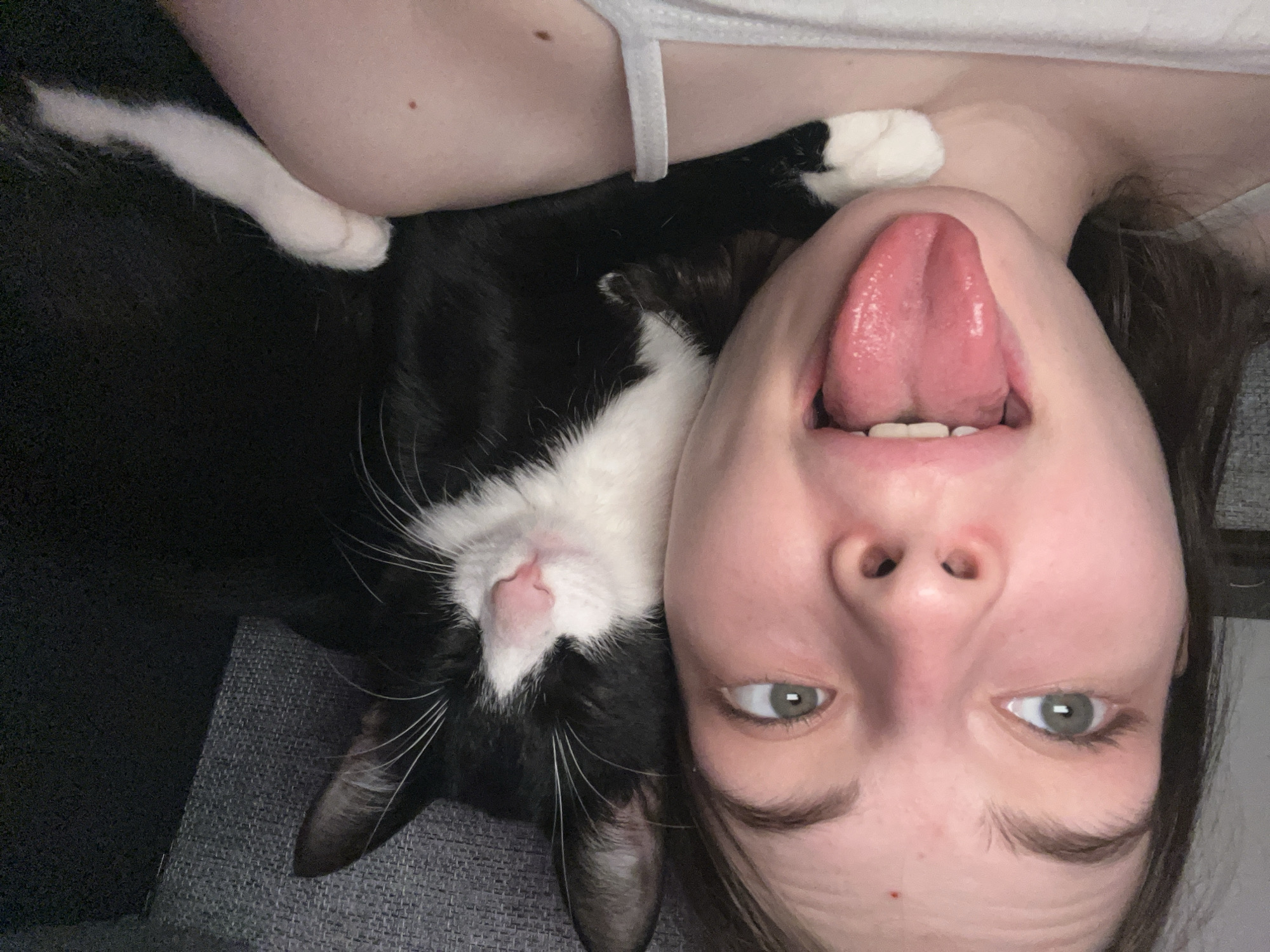 A close-up, upside-down selfie shows a young woman with light skin and green eyes, sticking her tongue out and looking directly at the camera. A black and white tuxedo cat is nestled near her neck, with its eyes closed. The background is a neutral gray, appearing to be a piece of furniture. There is no evidence to suggest the location is Saint Petersburg, Russia.