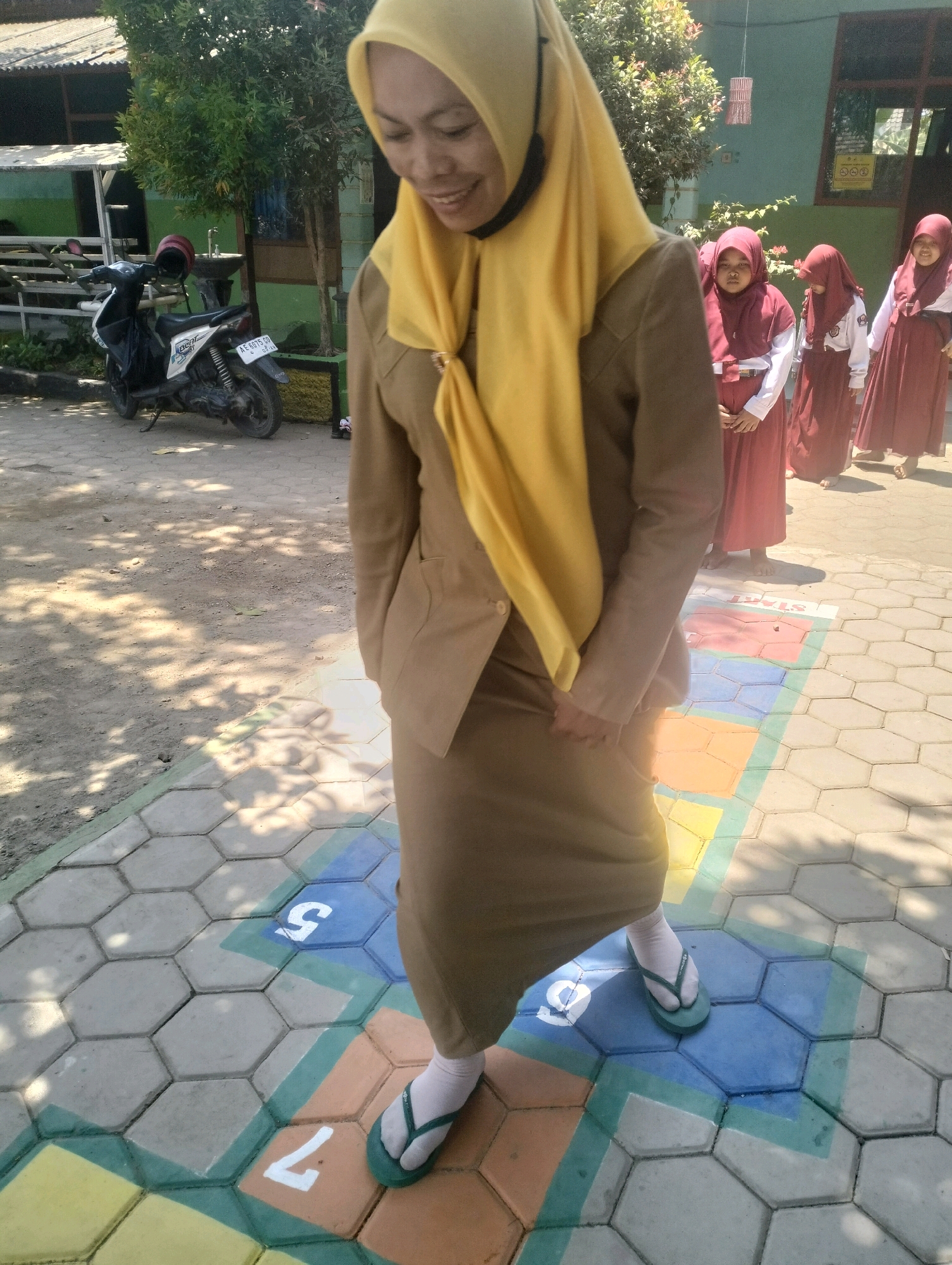 A woman wearing a tan uniform and a yellow hijab, smiles as she walks on a hopscotch court made of hexagonal paving stones painted with colored squares and numbers. She is wearing white socks and green flip-flops. In the background, three young girls in maroon hijabs and white tops stand on a paved area. A motorcycle is parked to the left of the frame, near a low structure with a green wall and a roof. The scene appears to be outdoors, possibly at a school in Blabakan, Indonesia, given the presence of children in uniform and the architectural style. The ground is dappled with sunlight and shadows, suggesting a bright, sunny day.