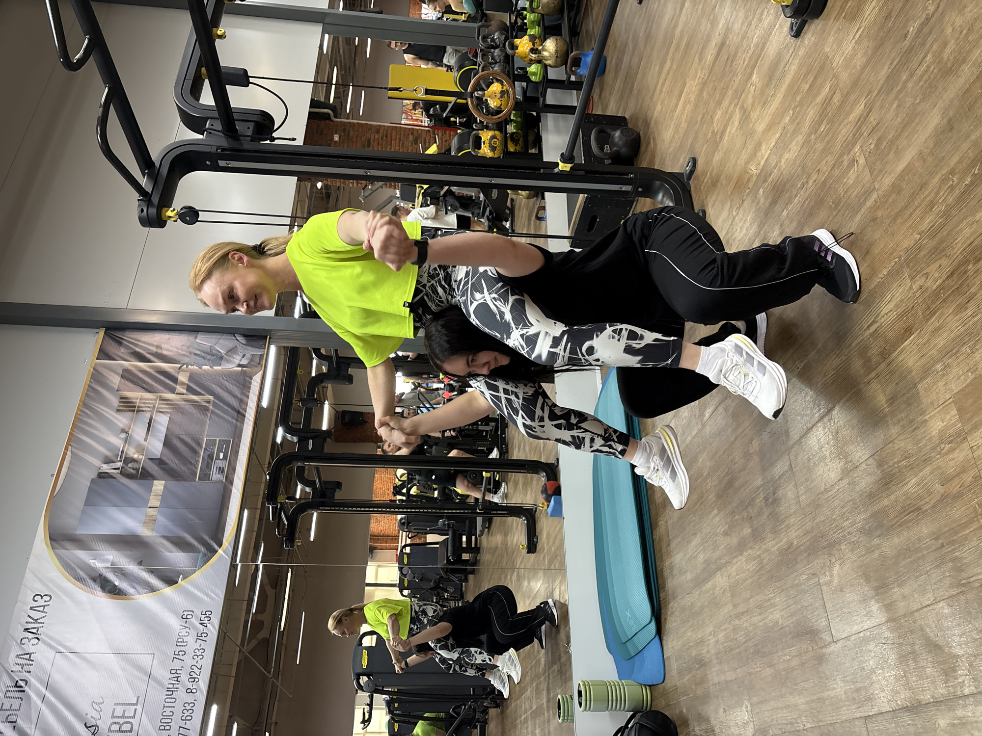 The image displays two women exercising in an indoor gym, captured from a slightly elevated perspective. The photograph is rotated 90 degrees clockwise.
**Subjects:** A woman with blonde hair, tied back, wears a bright neon yellow short-sleeved top and black-and-white patterned leggings. She is positioned to provide support, holding the hands or wrists of a second woman. The second woman, with dark hair, wears a black long-sleeved top and black track pants over similar black-and-white patterned leggings. She is horizontally extended, with her feet off the ground, performing a partner plank or "superman" exercise, suspended by the first woman's grip. Both women are wearing athletic sneakers.
**Setting:** The gym features a light wooden floor and various black metal gym equipment, including a cable machine and a rack of kettlebells, visible in the background. A large mirror on the left reflects the women and the gym space, showing their mirrored images. A blue exercise mat and a green foam roller are on the floor near the mirror. The gym is brightly lit by overhead artificial lighting, and parts of the walls appear to be grey or exposed brick.
**Notable details:** The women appear focused on their exercise, demonstrating teamwork. The environment suggests a dedicated fitness facility.
**Visible text:** A large banner, primarily visible in the mirror's reflection, displays text in Russian. It reads "МЕБЕЛЬ НА ЗАКАЗ" (CUSTOM FURNITURE), followed by "sia BEL," an address "ВОСТОЧНАЯ, 75 (РСУ-6)" (VOSTOCHNAYA, 75 (RSU-6)), and two phone numbers: "77-633" and "8-922-33-75-455".