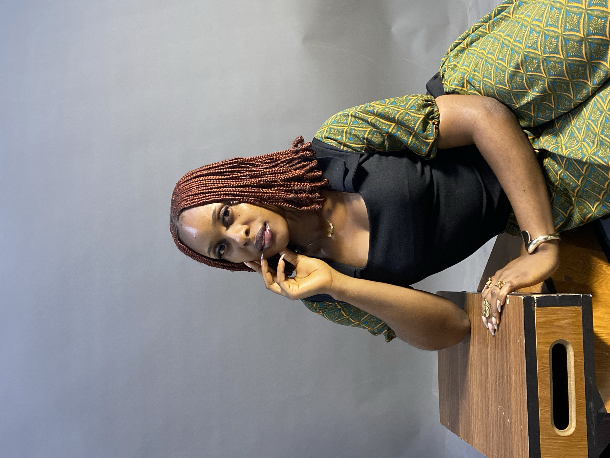 A woman is seated, leaning on a wooden box with an opening on its side. She has reddish-brown braided hair, a black top, and a green and yellow patterned dress with short puffy sleeves. She wears gold jewelry, including rings and a bracelet. Her left hand rests on her cheek with her fingers extended upward. The background is a plain grey. The location is not explicitly identifiable as Jos, Nigeria, from the visual information alone.