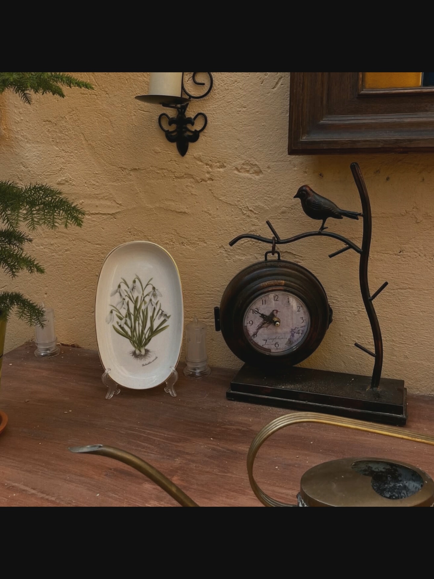 This is a medium shot of a wooden table with various decorative items on it, against a textured, cream-colored wall. On the left, a sprig of evergreen foliage is partially visible. Next to it, a white oval dish with a painted floral design sits on a clear stand. Two small, clear glass candle holders are placed on either side of the dish. To the right of the dish is a dark, round clock with Roman numerals on its face, suspended from a decorative metal stand that resembles a tree branch. A small, dark bird figurine is perched on the top of the metal stand. The background wall has a framed painting or picture with a dark wooden frame. In the foreground, a portion of a brass watering can is visible, with its spout extending from the lower left towards the center of the frame. The overall scene appears to be an indoor setting, possibly a home or shop display, in Bielawska Wieś, Poland.