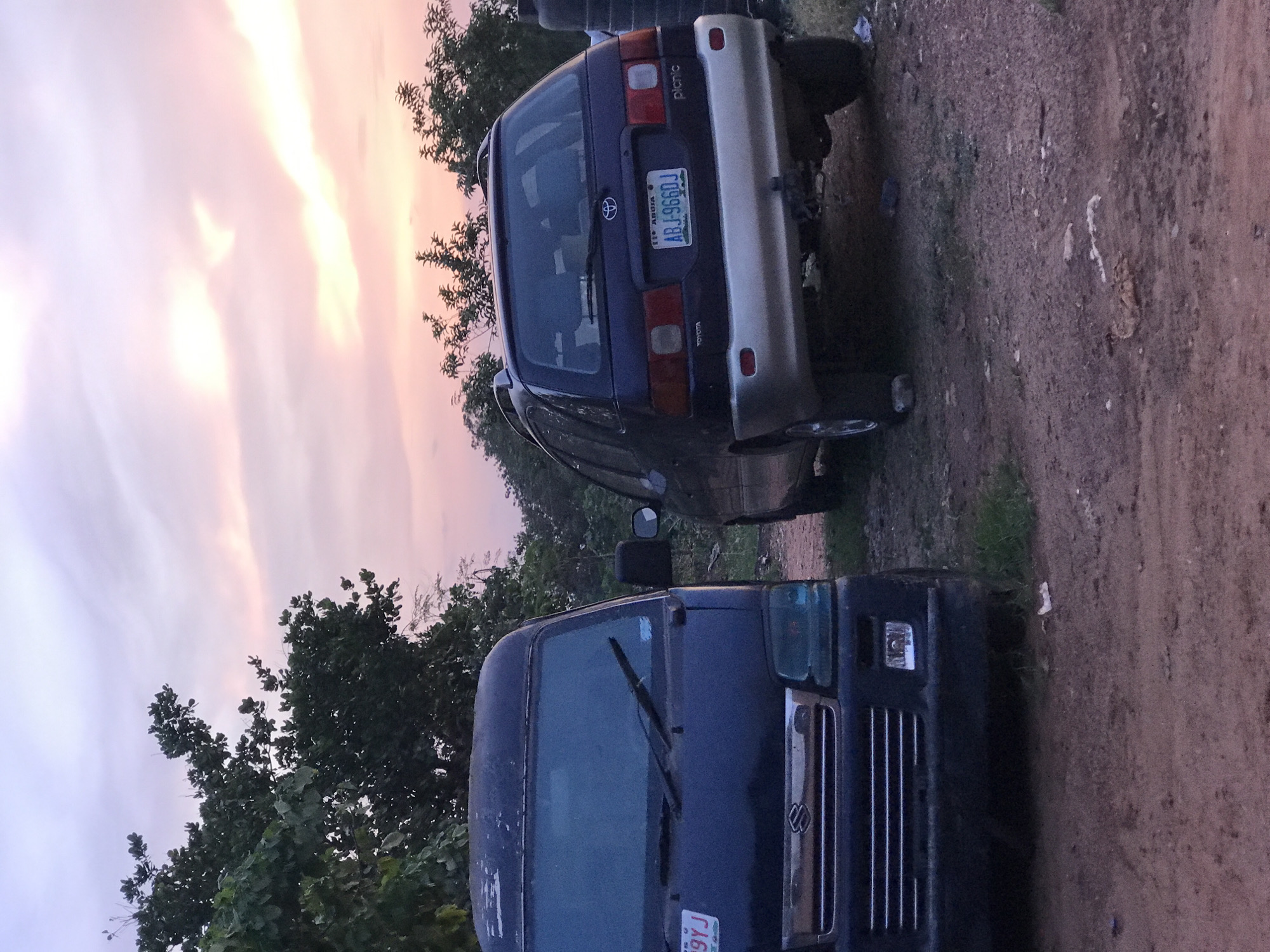 The image displays an outdoor scene in Galadimawa Village, Nigeria, likely during sunrise or sunset, characterized by a soft, colorful sky showcasing hues of pink, orange, and purple as light breaks through clouds. Two vehicles are parked on an unpaved, dirt ground.
In the background, the rear of a dark blue Toyota Picnic minivan is visible, featuring a prominent silver bumper. Text on the vehicle includes "Picnic" on the upper right and "TOYOTA" below the rear window. The Nigerian license plate clearly reads "FCT ABUJA" at the top, "NIGERIA" at the bottom, and the registration number "ABJ-986DJ" in the center.
Closer to the foreground and to the left, the front of a dark blue Suzuki minivan, possibly a Suzuki Carry, is partially visible. It has rectangular headlights, a chrome grille with the Suzuki logo, and a partial license plate ending in "99J".
Green trees and bushes flank the scene on the left and right, and in the distance, indicating a vegetated environment. The ground is unpaved and appears earthy. No people or specific activities are depicted; the vehicles appear to be stationary.