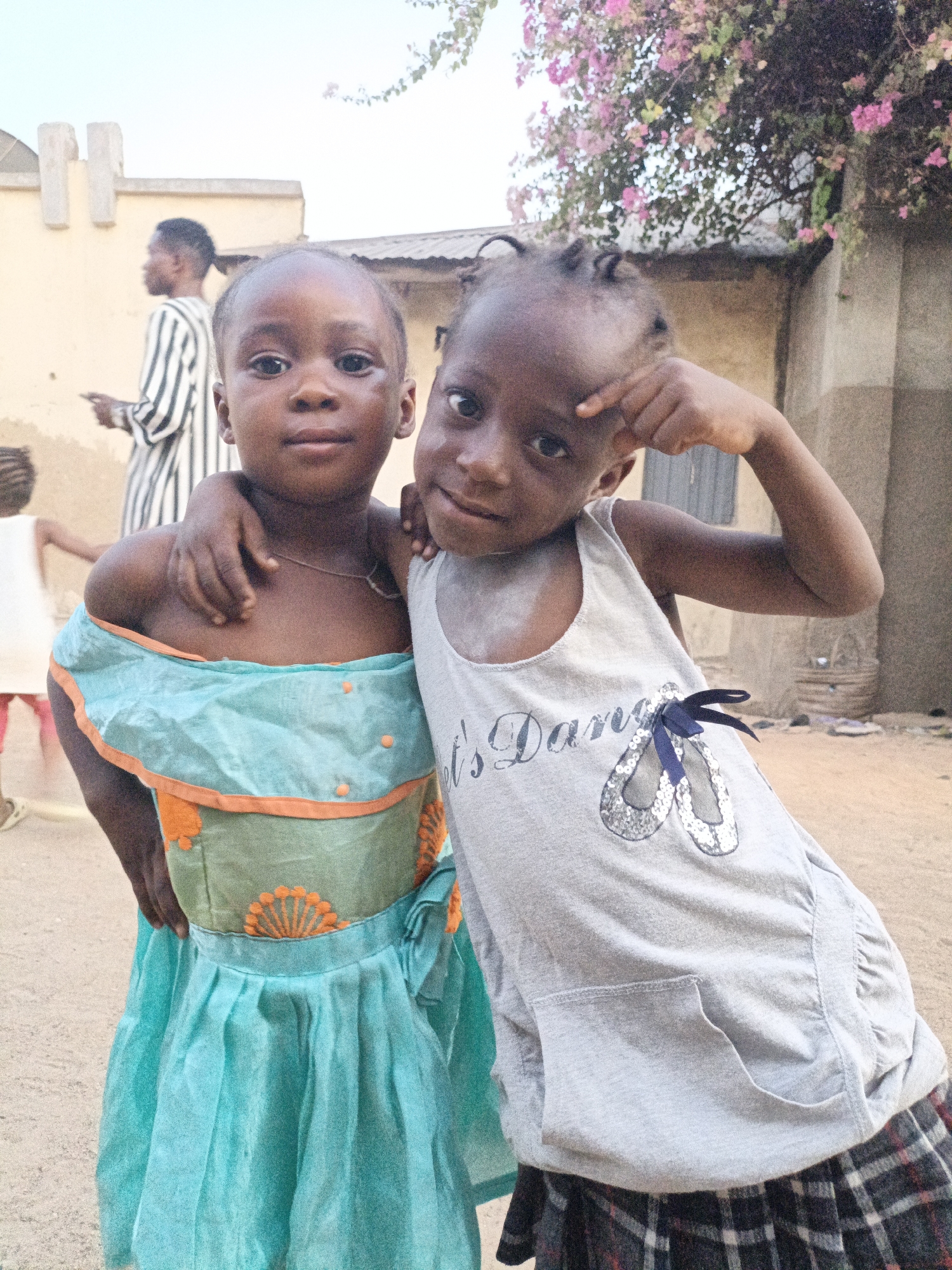 Two young Black girls stand outdoors. One wears a light blue dress with orange trim and floral accents, and the other wears a gray tank top with the words "Let's Dance" and a graphic of ballet slippers over a plaid skirt. The girl in the tank top points to her forehead. The girl in the dress has an arm around the other girl.
In the background is a partially visible building with two vertical, rectangular projections near the roof. A man in a striped shirt stands near the building. Pink flowers hang from a vine or tree near the right side of the building. A child in a white dress is walking near the foreground. The ground appears to be sandy. The location is Gwagwarwa, Nigeria.