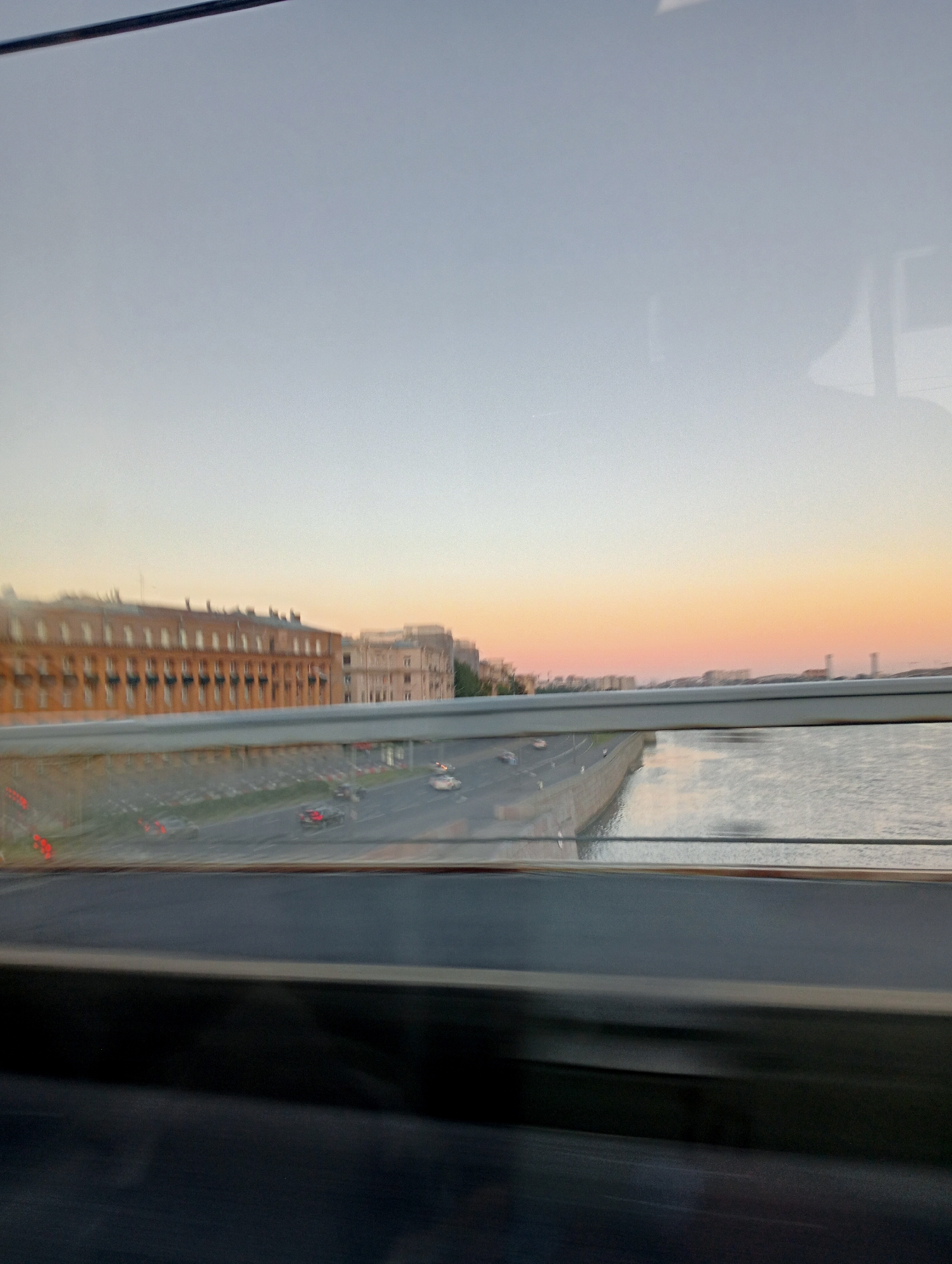 The image depicts a view from a bridge or elevated position looking out over a cityscape at sunset. The sky displays a gradient of colors, transitioning from a pale blue at the top to soft oranges and pinks near the horizon. In the distance, a river or body of water stretches across the scene, reflecting the sky. Along the far bank of the water, a row of buildings with distinct architectural features, possibly historical, are visible. Some of these buildings appear to be made of brick and have numerous windows. A road runs parallel to the river, with several cars in motion, their headlights and taillights blurred due to the motion of the camera or the vehicles themselves. A railing or barrier is prominent in the foreground, dividing the view of the road and water from the bottom portion of the image, which is dark and appears to be part of the structure from which the photo was taken, possibly a vehicle or another bridge. The overall impression is that of a cityscape at dusk, viewed during a journey, possibly by car or train.
