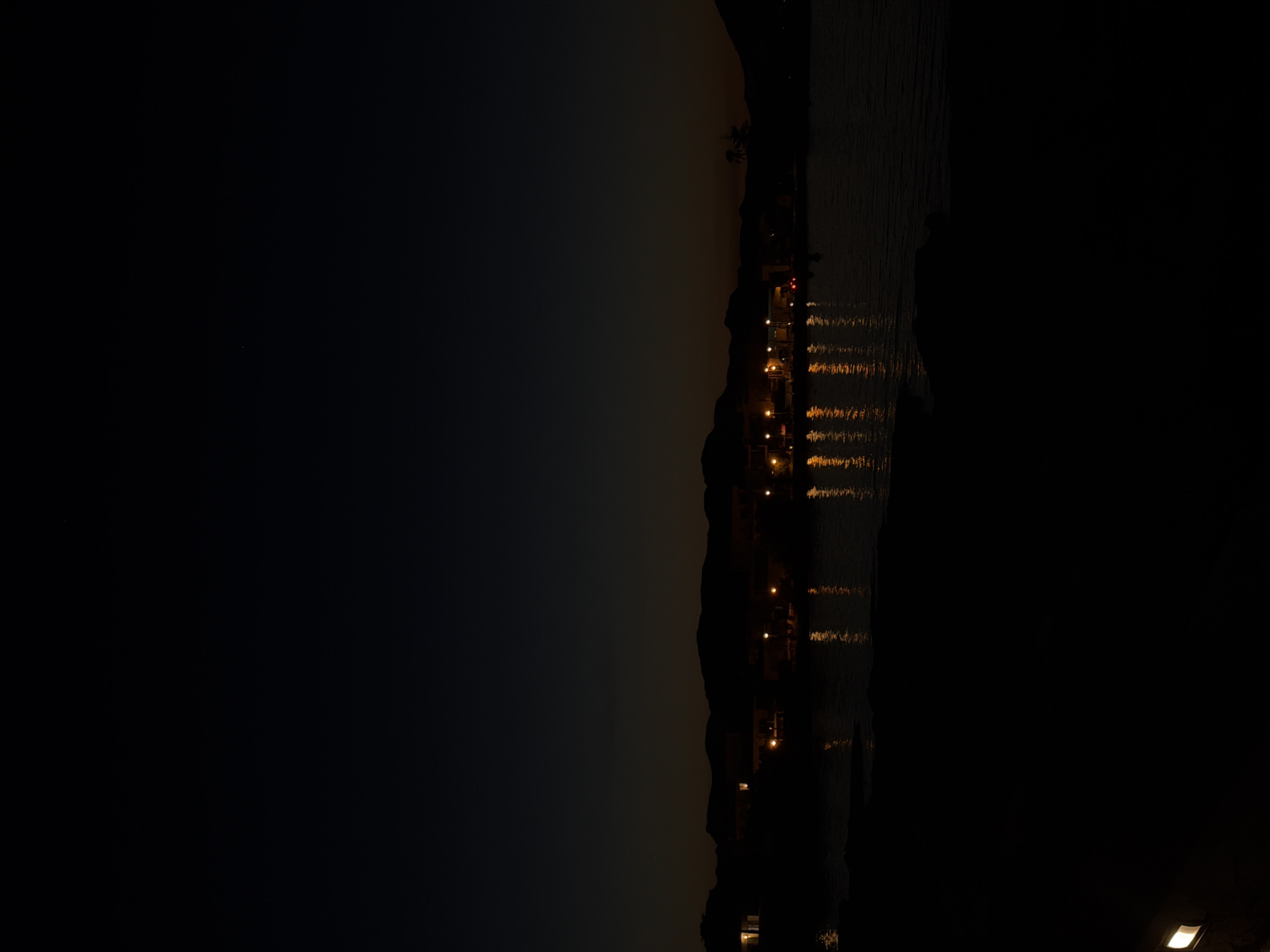 A nighttime scene in Alcúdia, Spain, shows a dark landscape bisected vertically. The right side consists of a body of water, possibly the sea, reflecting the yellow lights of buildings situated along the shore. The left side is dominated by darkness, suggesting a landmass or the night sky. The lit buildings appear to be arranged along a coastline, hinting at a populated area.