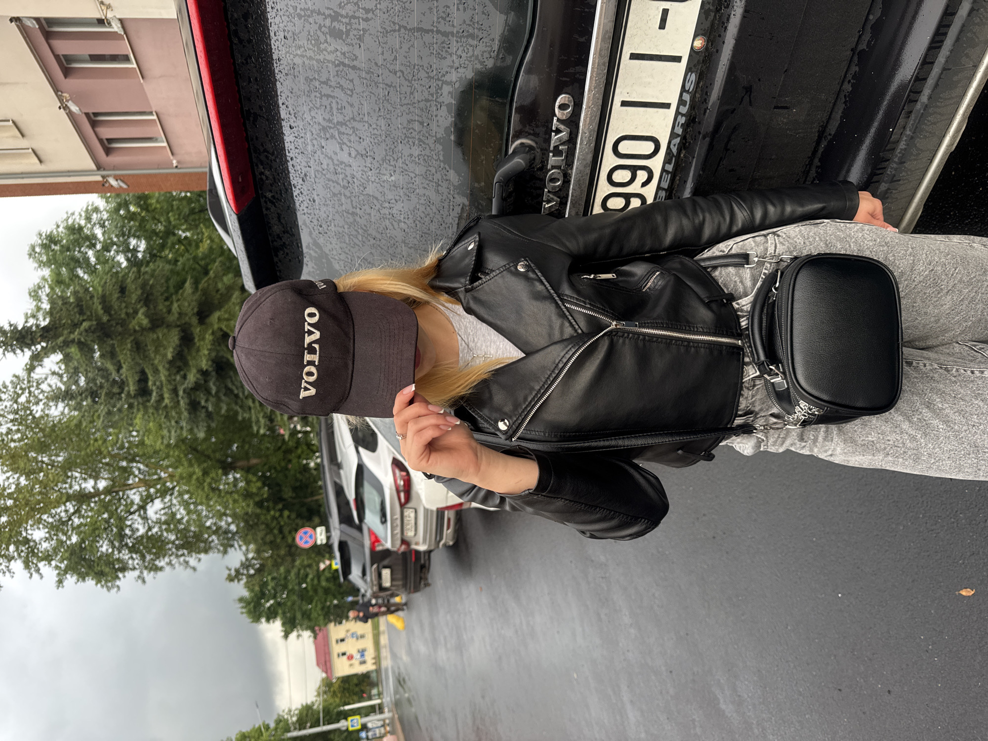 An individual is standing in front of a black Volvo SUV on a wet, paved street, likely in a city given the presence of parked cars and buildings in the background. The individual is wearing a black leather jacket, light-wash jeans, and a black baseball cap with the word "VOLVO" embroidered in white. They are also carrying a small black crossbody bag. The car's license plate is partially visible and reads "990 11-". The overall atmosphere appears to be overcast.
