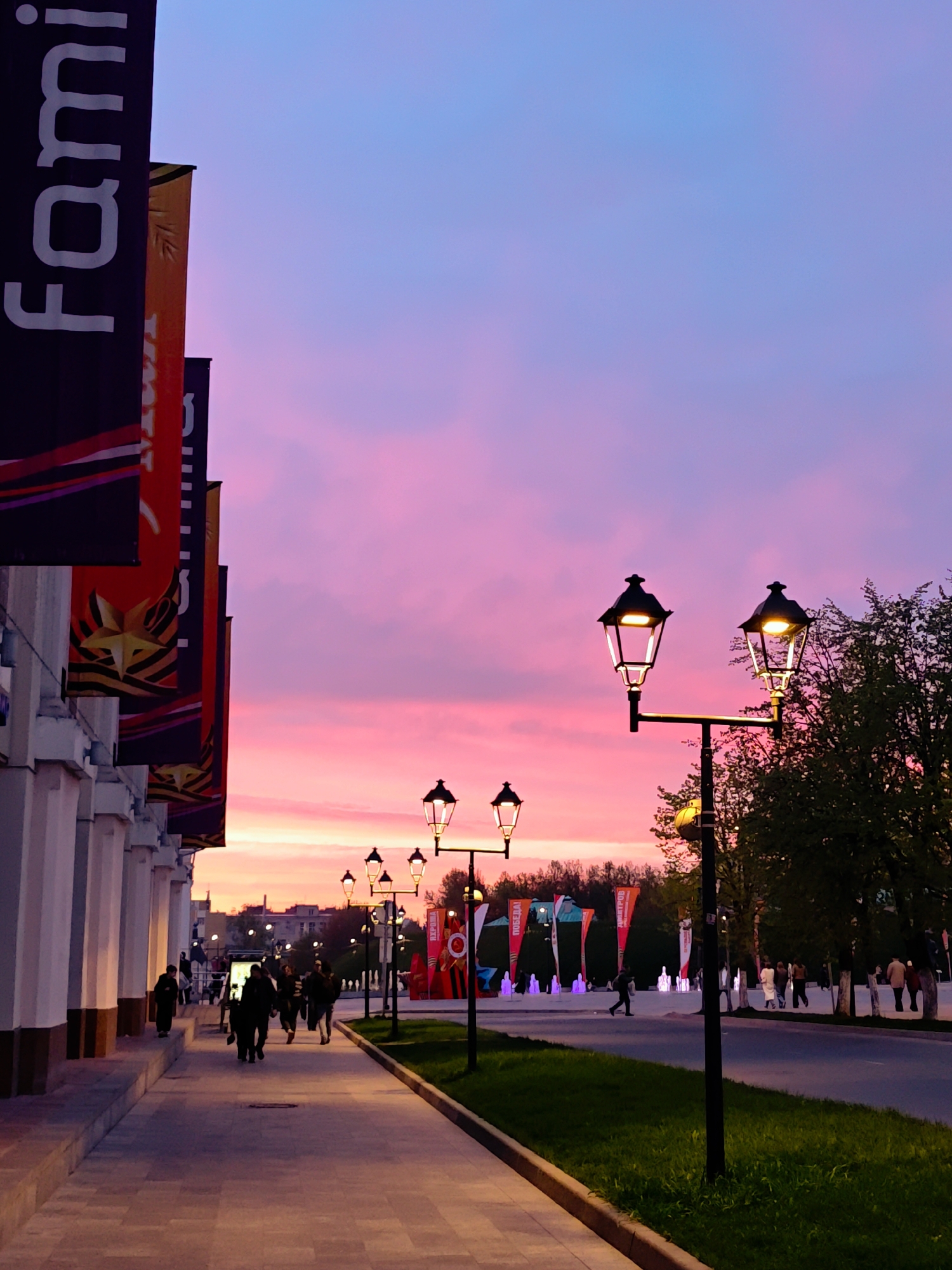 The image shows a street scene in Dmitrov, Russia at sunset. The sky is a mix of pink and blue hues. Along the left side of the image is a colonnade with banners hanging from it. On the right side is a line of street lamps, illuminated, and trees. People are walking on the sidewalk and in the street. In the distance, there are flags and fountains.