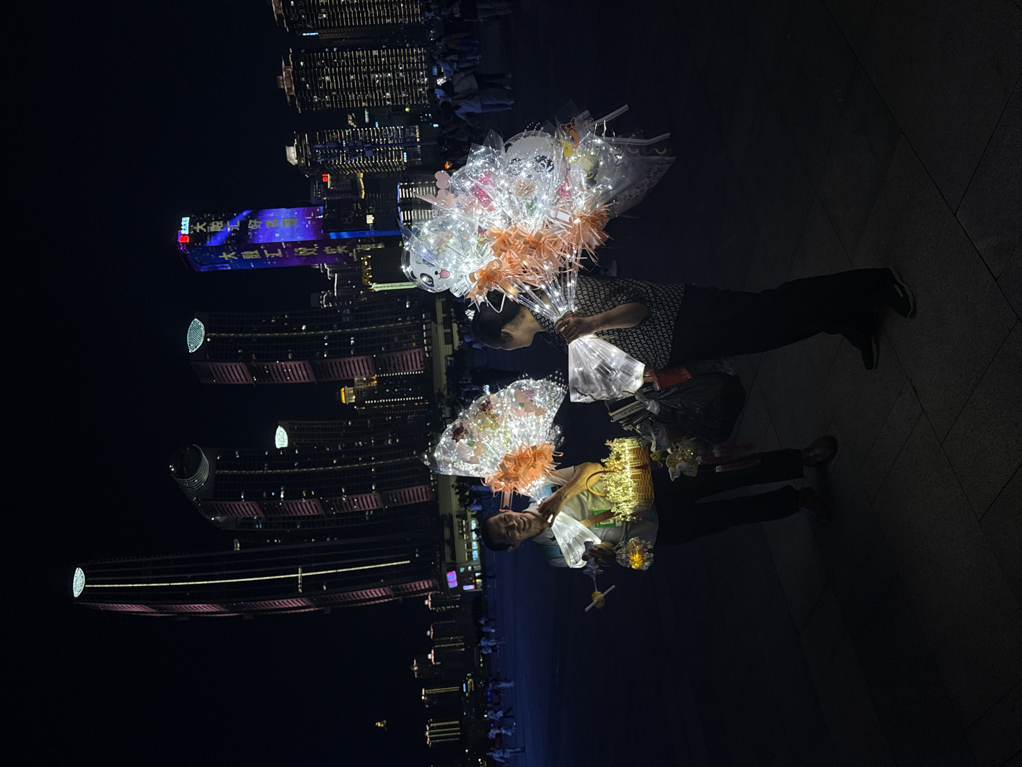 The image depicts a nighttime scene on a paved plaza in Jiangbeicheng Subdistrict, Chongqing, China, with several illuminated skyscrapers in the background. In the foreground, two vendors are selling illuminated decorative items, likely for the evening crowd. The vendor on the left, a man with a light-colored shirt and dark pants, is holding a basket filled with small, glowing ornaments and is showcasing a larger illuminated object. The vendor on the right, a woman wearing a patterned top and dark pants, is holding a large cluster of illuminated balloons and other decorative items, some of which resemble butterflies. Both vendors appear to be interacting with potential customers, though none are explicitly visible engaging in a transaction. The plaza is populated by many people strolling in the background, some of whom are also carrying illuminated objects. The skyscrapers are prominently lit with a combination of white, pink, and blue lights, some displaying text or patterns, creating a vibrant urban backdrop. The ground is dark and appears to be made of large, rectangular paving stones.
