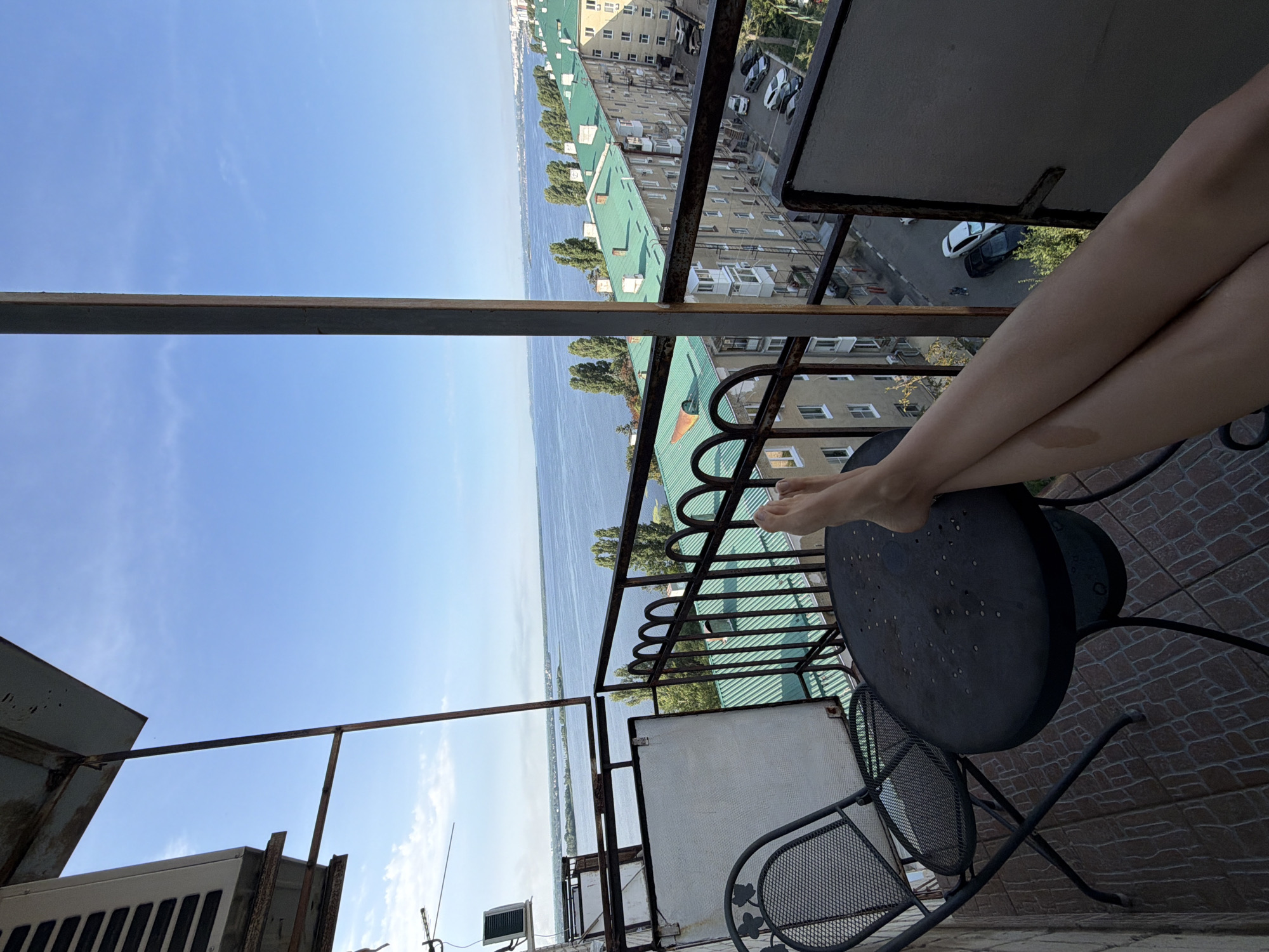 The image depicts a view from a balcony in Saratov, Russia, overlooking a wide river and a cityscape with apartment buildings. In the foreground, a woman's legs are visible, crossed and resting on a dark, round table. A black metal chair is to the left of the table, and the balcony floor is tiled with a brown, stone-like pattern.
The river dominates the mid-ground, with trees lining its banks and a distant shore visible. Beyond the river, a series of apartment buildings with distinctive green roofs stretch along the horizon, indicative of Soviet-era architecture. Cars are parked along a street running between the buildings. The sky is a clear, bright blue with a few wispy clouds. An air conditioning unit is mounted on the exterior wall of the building, and a metal railing with decorative arches encloses the balcony. The overall impression is one of a relaxed moment enjoying the view on a sunny day.