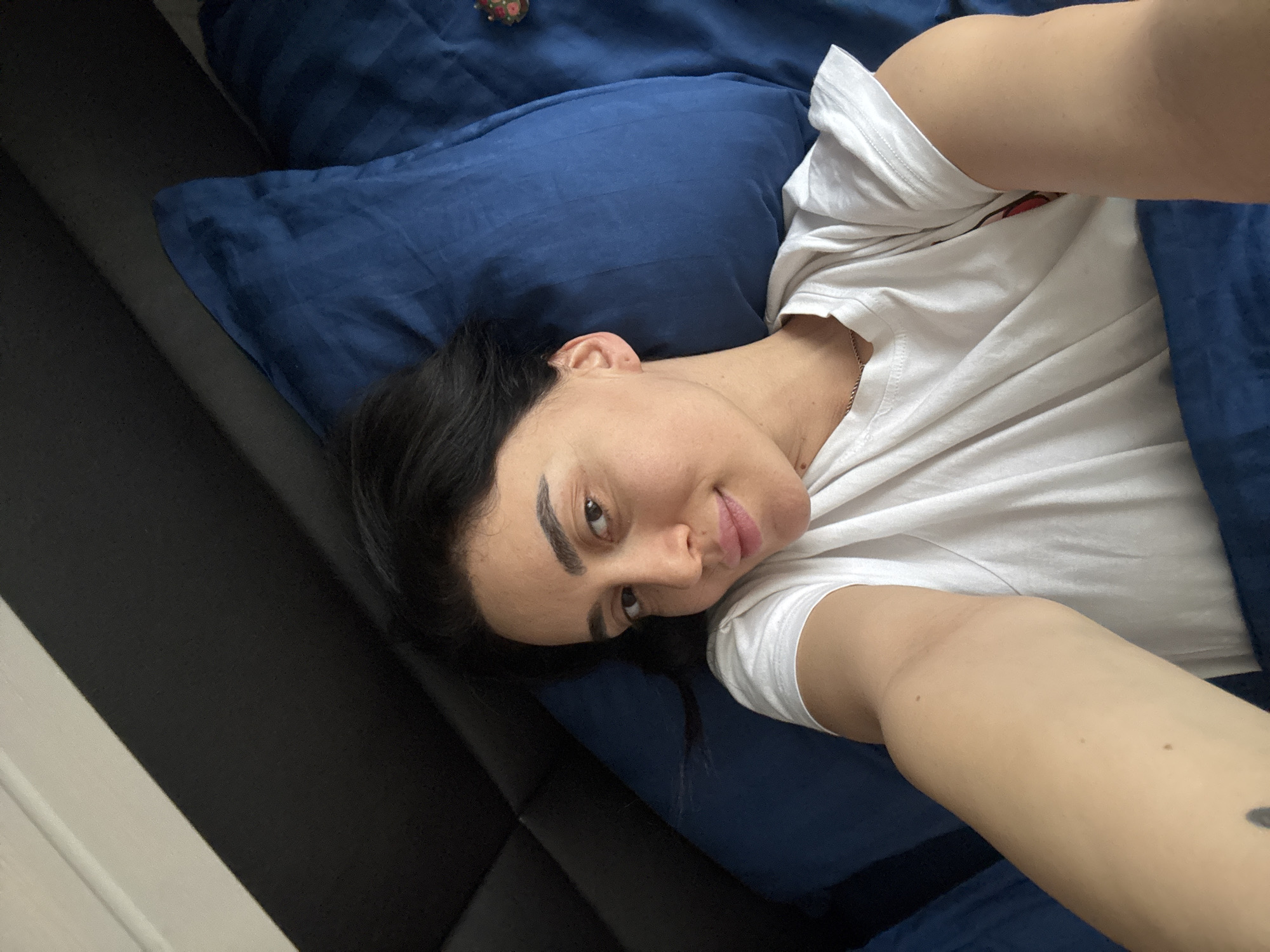 A young adult female is depicted in a selfie, lying on her back in a bed located in Tbilisi, Georgia. She has dark hair and eyebrows, with a light to medium complexion, and is looking directly at the camera with a subtle smile. She is wearing a white short-sleeved t-shirt and a thin, silver-colored chain necklace. Her right arm, visible in the foreground, is extended towards the camera, indicating she is holding the device. A dark mark, possibly a tattoo, is visible on her inner right upper arm. She is resting on a royal blue pillow, with a matching blue pillow to her right. A blue duvet or blanket covers her body from the chest downwards. Behind her head and to the upper left, a dark, possibly upholstered, headboard is visible. A small, multi-colored floral-patterned object rests on the blue pillow to her right. The scene is illuminated by soft, even lighting, consistent with an indoor bedroom environment.