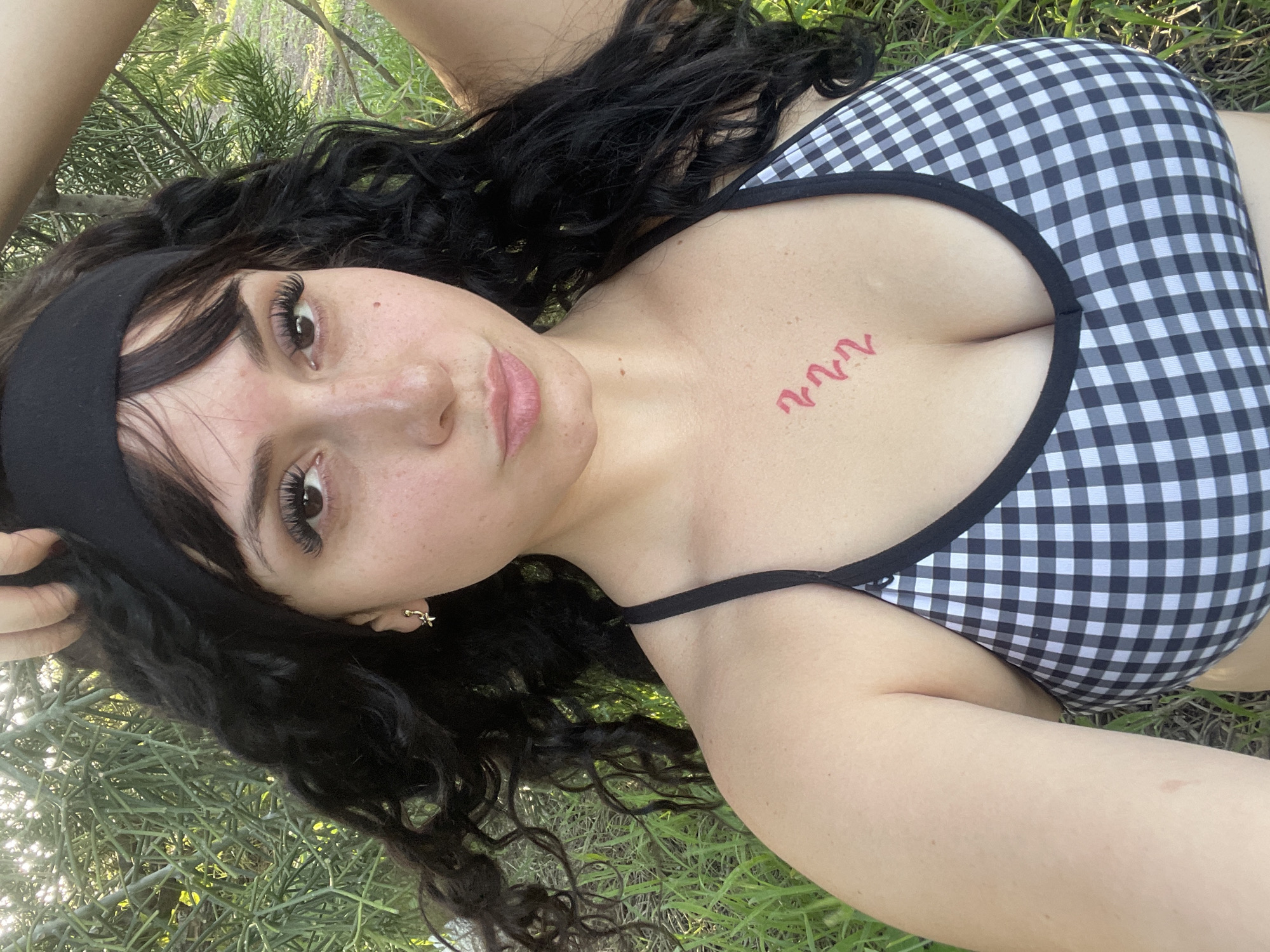 A young woman with long, dark, curly hair and bangs, wearing a black headband and a black and white gingham bikini top, is outdoors. She has a small, red, wavy tattoo on her chest. She is looking towards the camera with her head tilted slightly back, and her left hand is resting on top of her head. The background consists of green foliage and grass, suggesting a natural outdoor setting. The lighting indicates it is daytime, with sunlight filtering through the leaves. There is no discernible location in the city of Maracay, Venezuela.