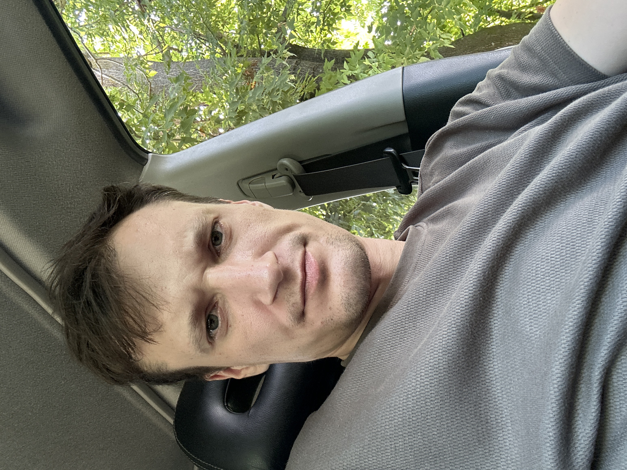 A man is seen from the chest up, inside a car. He has brown hair and is looking towards the camera with a neutral expression. He is wearing a grey t-shirt. The car interior is visible behind him, with the headrest of a black seat and a grey seatbelt. Outside the car window, trees and green foliage are visible, suggesting a natural or park-like setting. The light suggests it is daytime. There are no visible signs indicating the specific location within the city of Makiv, Ukraine, beyond the general context of being in a car in a green area.
