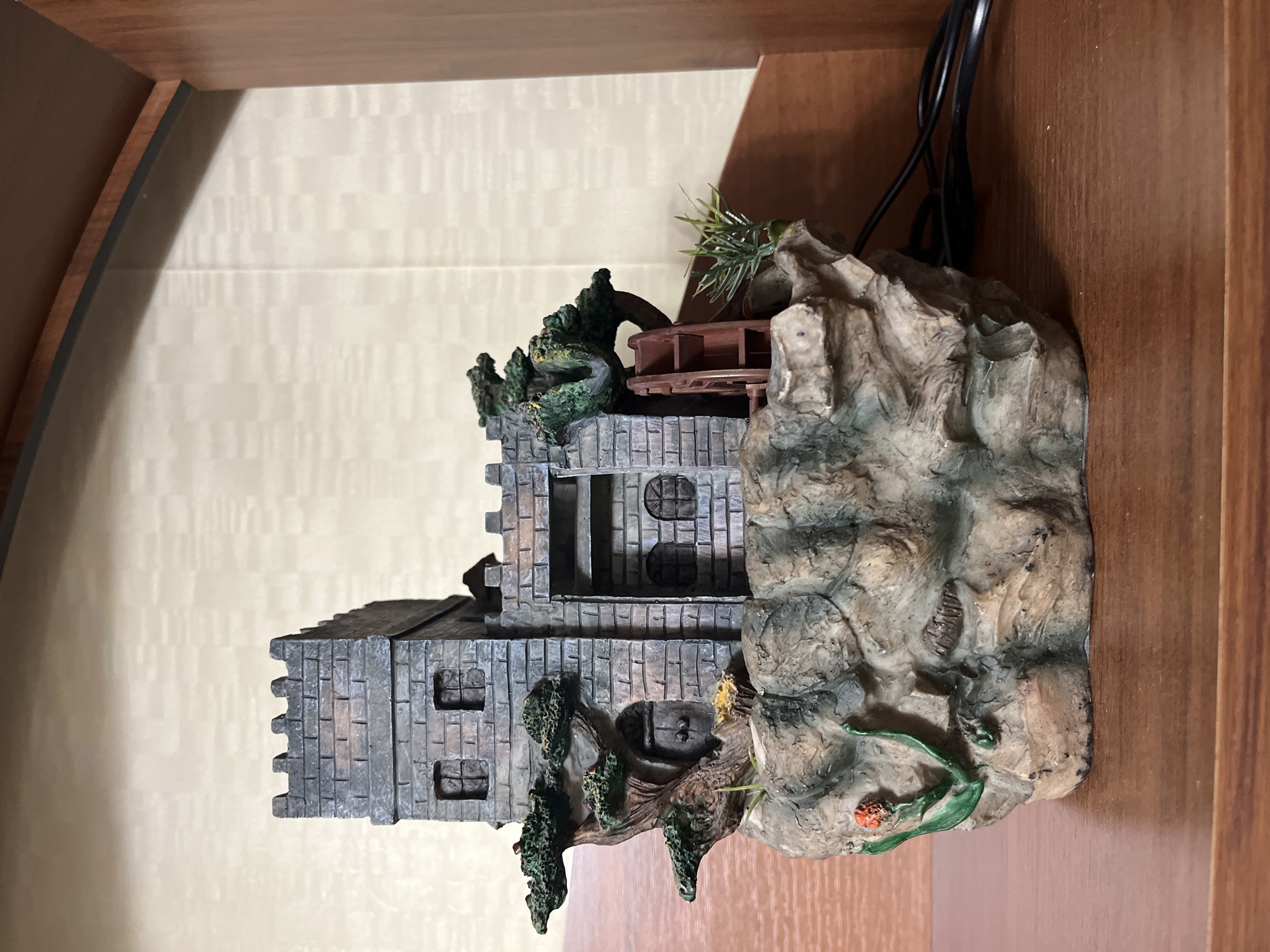 A detailed miniature structure, resembling a castle or fortress, sits on a warm brown wooden surface. The structure is built with textured, block-like walls in shades of gray, mimicking stone masonry, complete with battlements at the top and several small rectangular and circular window openings. Green, moss-like foliage and small trees are integrated around the base and sides of the castle, suggesting an aged or overgrown appearance.
Adjacent to the castle is a rugged, rocky formation rendered in various tones of gray, brown, and green, which appears to be part of the same decorative piece. A small, dark red water wheel-like mechanism is visible nestled between the castle and the rocky outcrop. Additional green foliage sprouts from the top of the rocky section, and a small reddish-orange flower detail is visible near the base of the rock.
Behind the entire setup, a light-colored wall with a subtle, vertical pattern serves as the background. Black electrical wires are visible extending upwards from behind the rocky formation, suggesting the object may be an illuminated decoration or a water fountain. The item appears to be an indoor decorative piece, possibly an aquarium ornament or a tabletop fountain. No human figures or discernible activities are present, and there is no visible text.