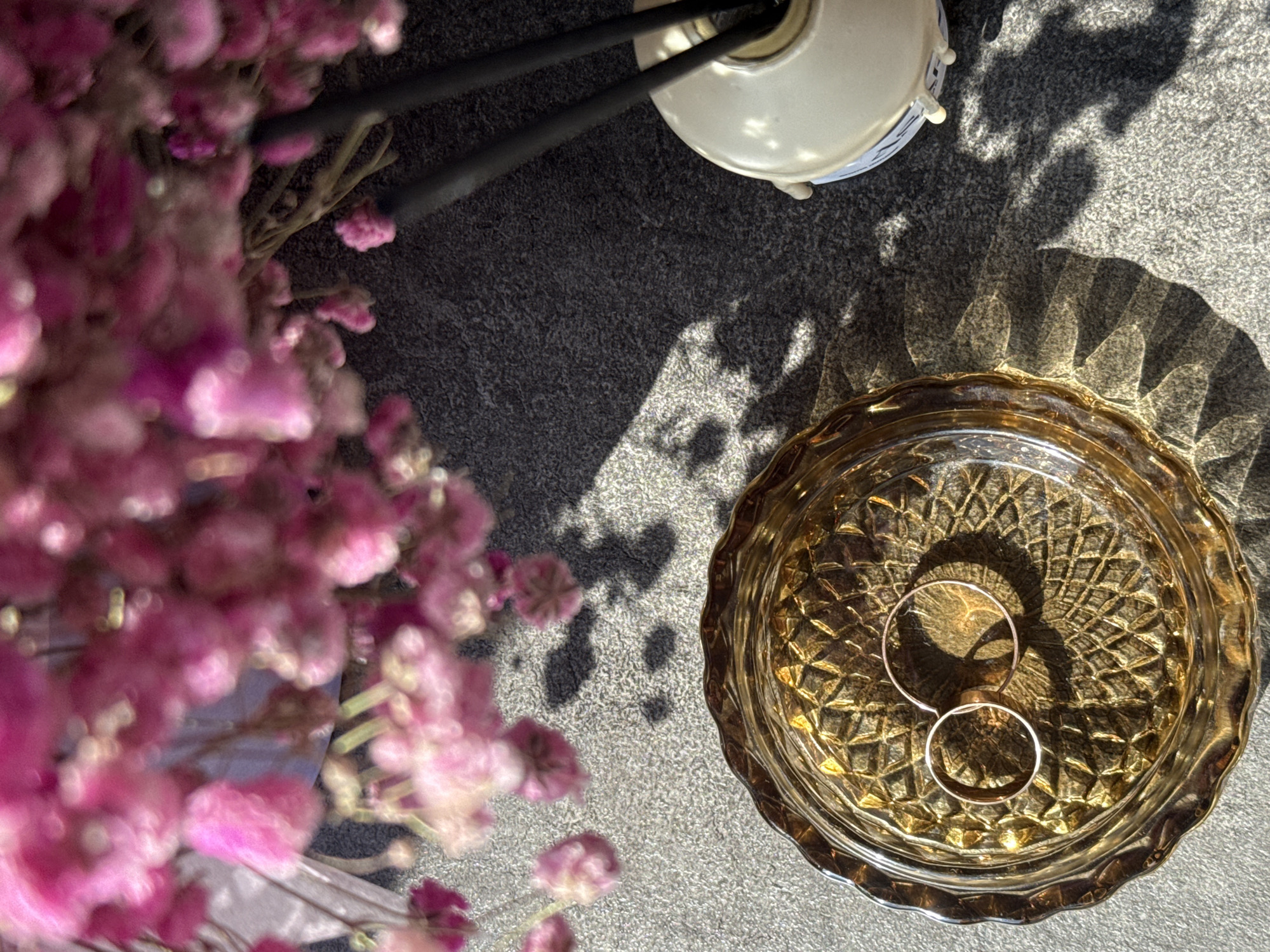 A high-angle shot captures two gold rings resting in a cut-glass dish, dappled with light and shadow. The dish is positioned on a textured, neutral-colored surface. In the periphery, soft pink flowers blur on the left, while parts of a lamp base are visible above the dish. The setting could be indoors, given the contained arrangement of objects. This image does not depict any discernible location in Petropavlovsk-Kamchatsky, Russia.