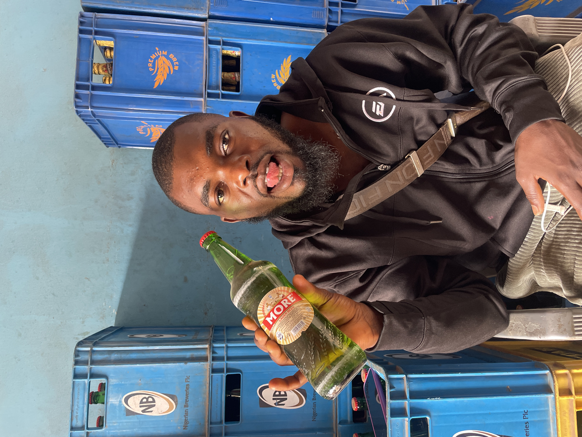 The image shows a man holding a bottle of "MORE Lager Beer" with a red cap, in front of stacked blue plastic crates labeled "Nigerian Breweries Plc." Some crates feature "Premium Brew" and "Trophy" branding. The man is wearing a black hooded sweatshirt with a white logo and a strap across his chest, sticking his tongue out in a playful expression. The background appears to be an indoor space, possibly a storage area, with light blue walls. The image does not contain any information regarding the city of Jos, Nigeria.