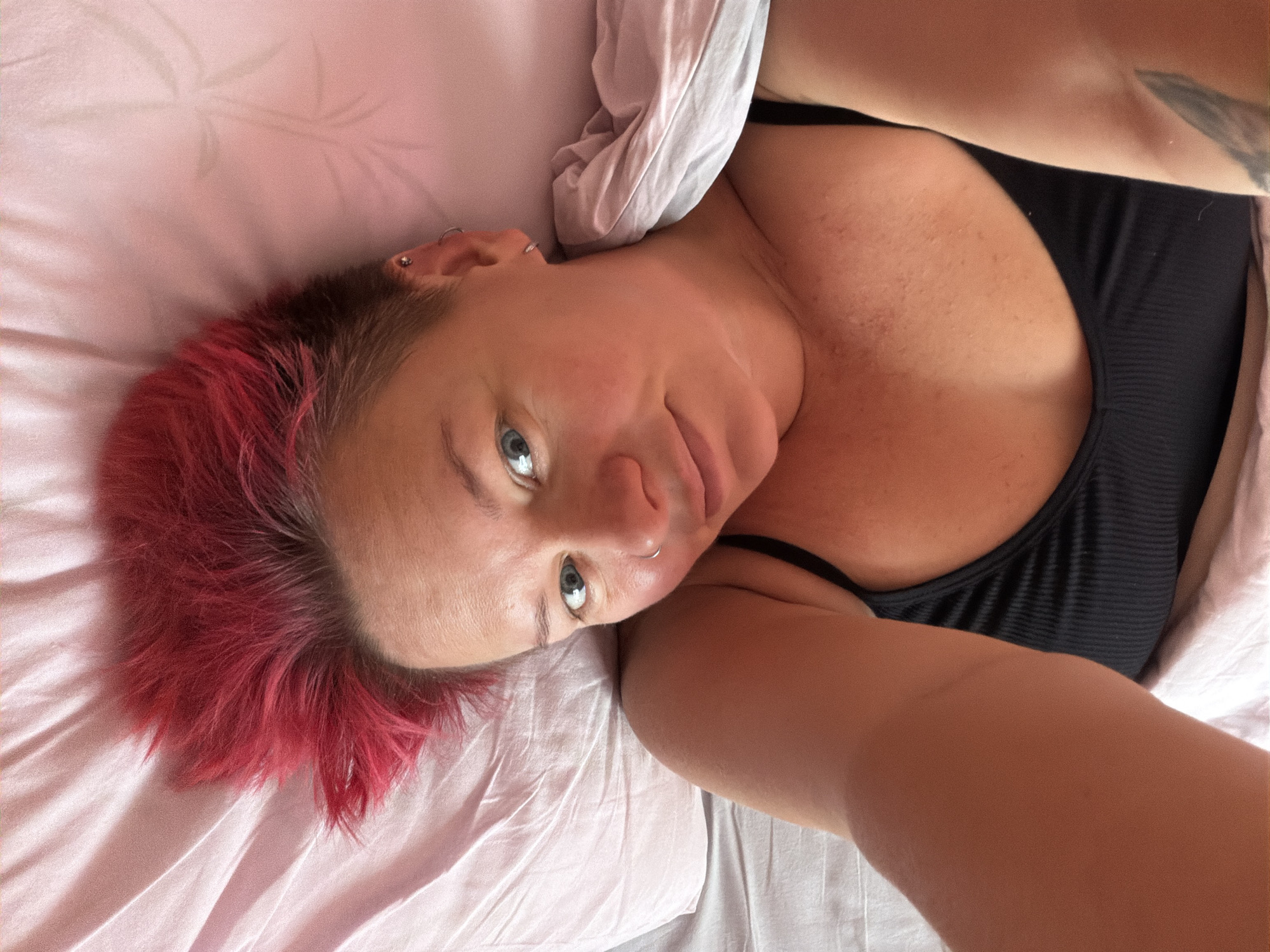 A young woman with bright pink, spiky hair lies on a bed with pink sheets. She is wearing a black ribbed tank top. She is looking at the camera with blue eyes and has a nose ring and an earring. A duvet is partially covering her. The scene is set in a bedroom in the city of Chaikovsky, Russia.