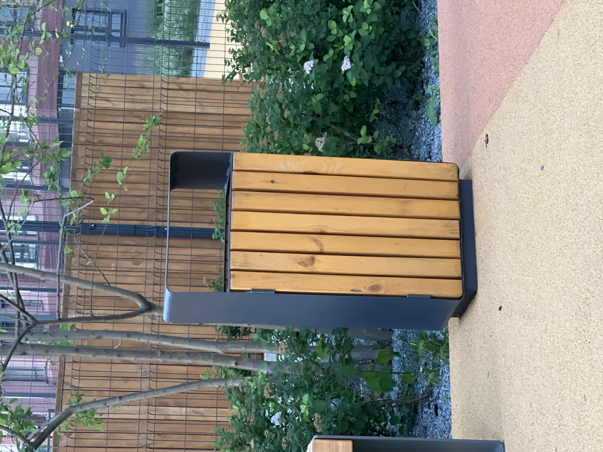 The image shows a modern, slatted trash bin with a dark grey metal frame and light wood slats. It is situated on a speckled, multi-colored paved surface, with a section of yellowish-tan material on the left and a section of reddish-pink material on the right. Behind the trash bin, there is a dense green bush with small white flowers. A vertical wooden fence, made of closely spaced planks, runs behind the bush and extends to the left. In front of the fence and behind the bush, several thin tree trunks are visible. Beyond the fence, a dark metal mesh fence encloses an area with buildings and more foliage. The scene appears to be an outdoor public space, possibly a park or a courtyard in a residential complex, in Yekaterinburg, Russia.