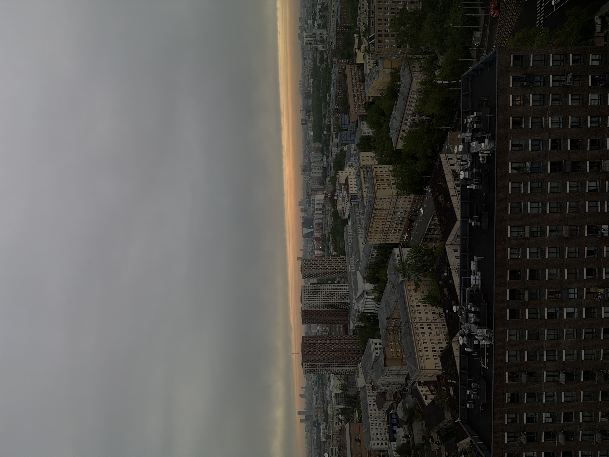 The image shows an aerial view of an urban area at dusk or dawn. The scene is divided into three main sections: the sky, the city skyline, and foreground buildings. The sky is mostly covered by grey clouds with a band of orange/yellow light on the horizon, suggestive of sunrise or sunset. The city skyline is a mix of residential and commercial buildings of varying heights and architectural styles. The foreground features a tall brick building.