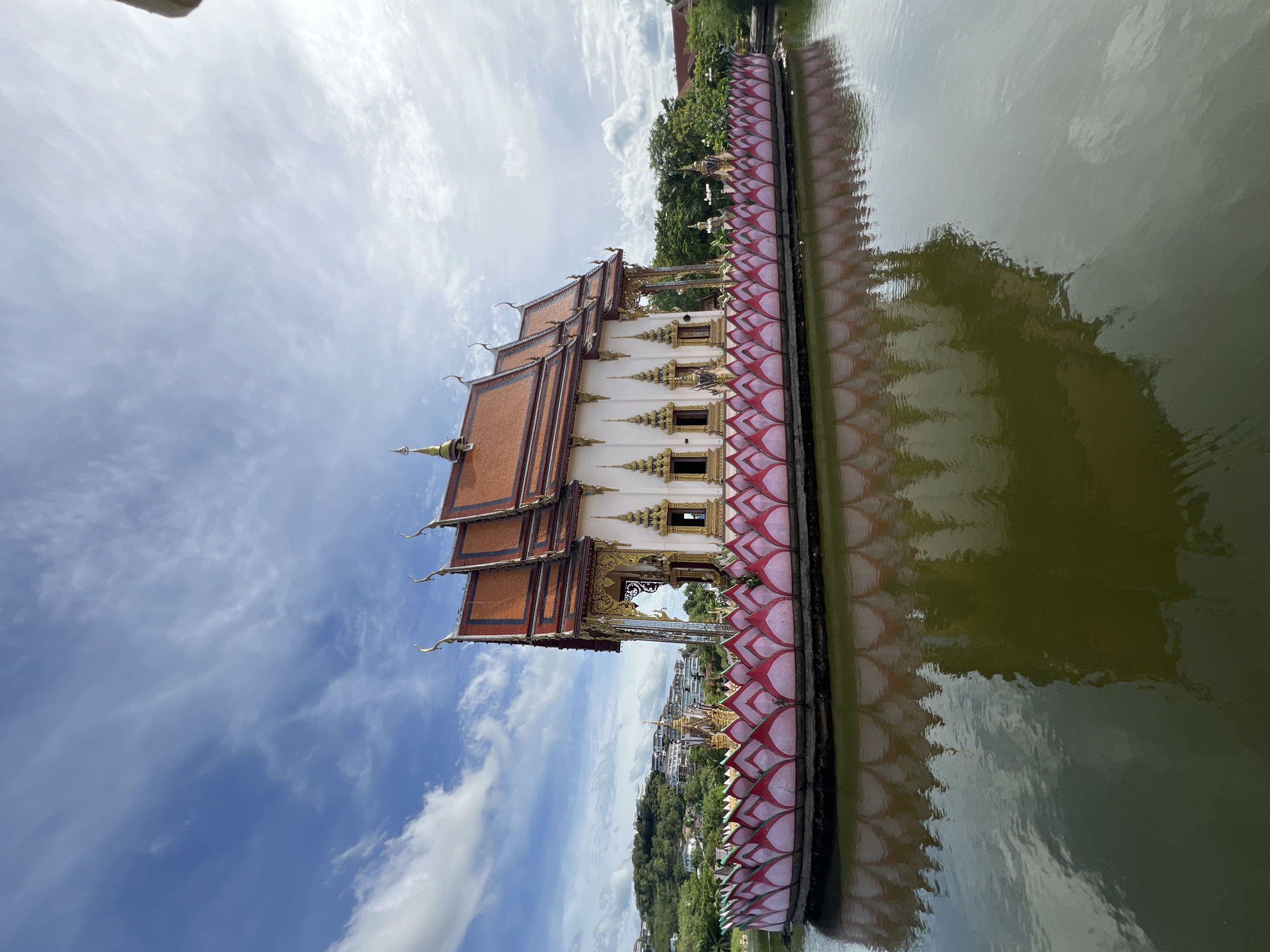 A Thai temple with an orange tiled roof and white walls sits on a circular platform surrounded by a pink and white lotus petal-like barrier in the middle of a body of water. The temple has ornate golden decorations around the windows and roofline. The water reflects the temple and the lotus barrier. In the background, a green, tree-covered hillside is dotted with buildings and the sky is blue with scattered white clouds. The scene appears to be in the Ko Samui District of Thailand. There are no people or animals visible in the image.