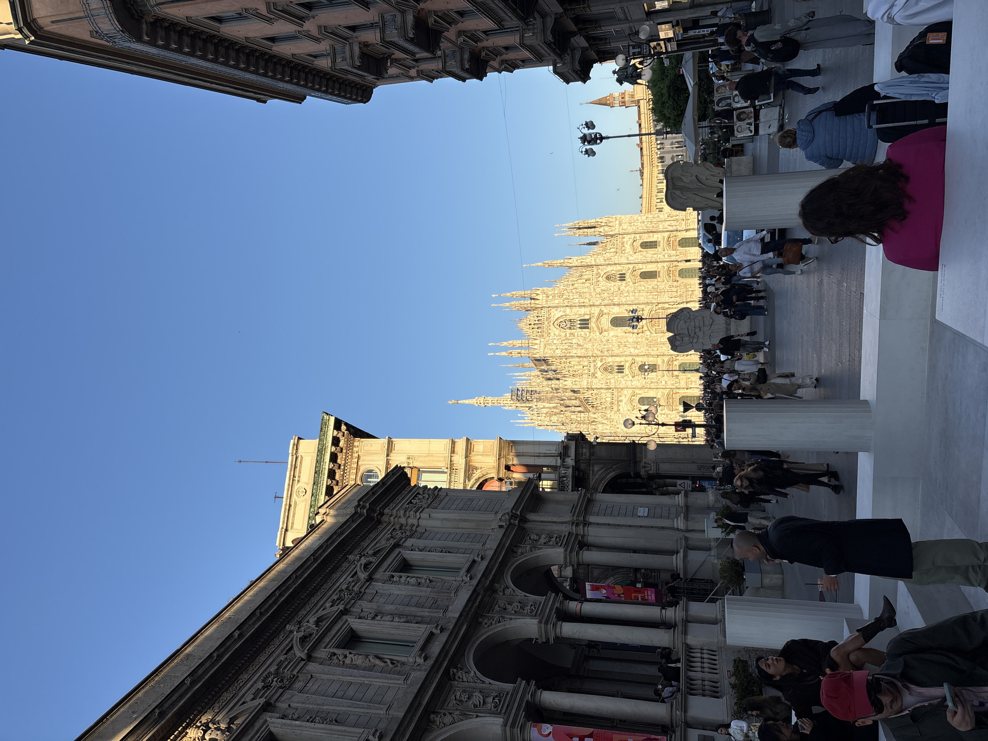 The image presents a vibrant, sunny scene of Milan's Piazza del Duomo. When viewed in its correct orientation, the magnificent Milan Cathedral (Duomo di Milano) stands as the dominant subject in the background, its intricate Gothic facade and numerous spires bathed in warm, golden light under a clear, bright blue sky.
In the foreground and to the left, a grand, multi-story building featuring classical architecture with detailed columns, arches, decorative carvings, and balconies frames the lower left portion of the image. On the ground level of this building, colorful banners are visible, with "CULTURA MILANO" partially discernible on one. To the right, various people are seated on modern, stark white tiered structures or benches, facing towards the cathedral or observing the plaza. One person in a bright pink top is clearly visible from behind, along with another wearing a red cap.
The plaza area is bustling with activity, as numerous individuals walk and gather. Some appear to be tourists sightseeing, while others are simply moving through the public space. In the mid-ground, there are further crowds near the cathedral, and what appear to be street vendors with displays can be seen on the right. Distant buildings and a distinct bell tower are visible beyond the main cathedral, further emphasizing the urban setting. The overall impression is that of a lively public square on a beautiful, sunny day, likely in the late afternoon or early evening, given the quality of light.