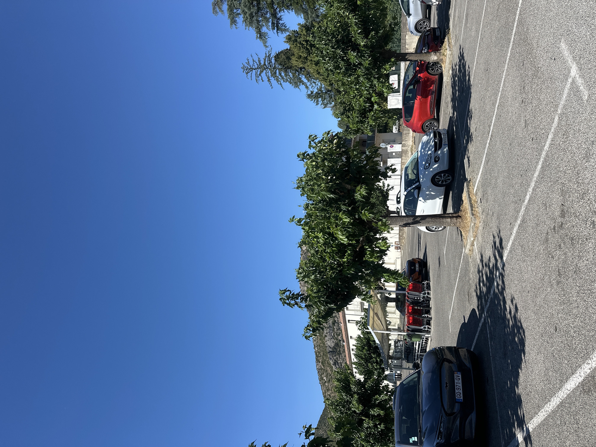 In Anduze, France, this image captures a sunny day in a parking lot with a clear blue sky. Several cars are parked in designated spots, including a dark-colored electric vehicle in the foreground with a French license plate starting with "GQ". Further back, a white hatchback, a red hatchback, and a grey SUV are visible. Shopping carts are neatly arranged under a shaded canopy. Trees with lush green foliage are scattered throughout the lot, casting dappled shadows on the asphalt. In the background, a building with white walls and a terracotta roof stands against a backdrop of rocky, mountainous terrain. The scene suggests a typical day in a French town, likely near a commercial area or a residential building.
