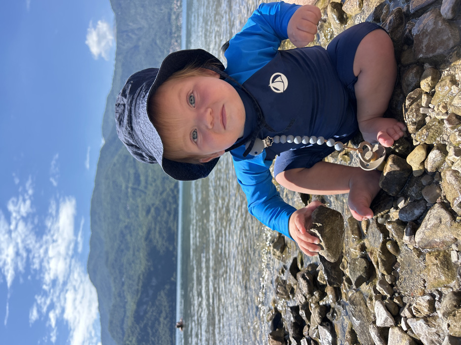An infant is positioned in a low squat on a rocky shoreline, facing directly towards the camera. The infant has light-colored hair, blue eyes, and rosy cheeks, and is wearing a navy blue bucket hat, a two-toned blue long-sleeved rash guard featuring a white sailboat logo on the chest, and navy blue swim shorts. A grey beaded pacifier clip, with a grey animal-shaped attachment, is clipped to the rash guard and extends to a light brown pacifier resting near the infant's left foot. The infant's left hand is grasping a grey rock. The immediate foreground and midground consist of a wet, rocky beach and shallow, clear water. A large body of water, consistent with Lake Como, stretches behind the infant. In the far left background, a person's head and shoulders are visible above the water line. Across the lake, steep, forested mountains rise prominently. The sky above is blue with scattered white clouds. The scene is illuminated by direct sunlight, indicating a clear day in Abbadia Lariana, Italy.