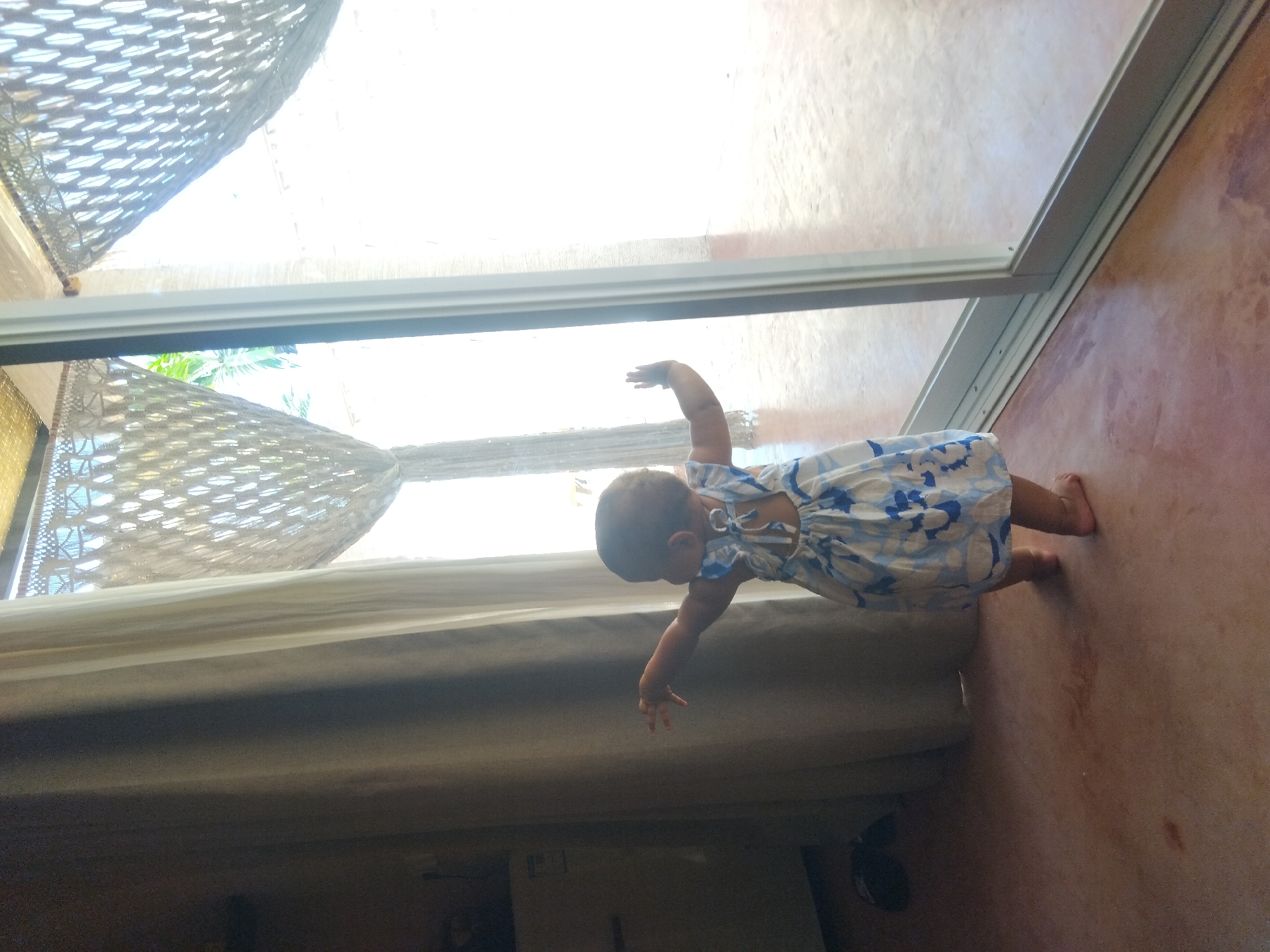 A baby wearing a blue and white patterned dress is standing near a sliding glass door. The baby is reaching toward the door with both hands, with one hand extended higher than the other. Behind the baby, there is a hammock hanging inside the room. The floor on which the baby is standing appears to be a reddish-brown color. The location of the scene is Unknown, Unknown.