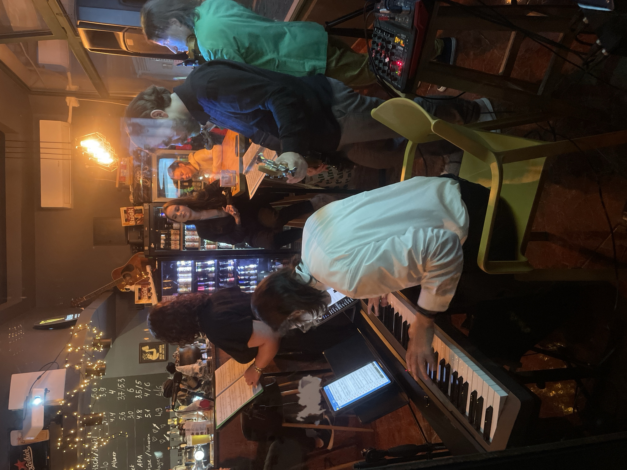 The image shows a band performing in what appears to be a bar or pub. There are at least five people visible: one playing a keyboard, one playing a guitar, another manipulating a sound mixing board, and two others, possibly singers or instrumentalists. The bar area includes beer price list on chalkboard, liquor bottles, and general bar paraphernalia. The band is illuminated by dim, warm lighting from a mix of decorative string lights and other fixtures. The location, Budva, Montenegro, is deduced from signs.