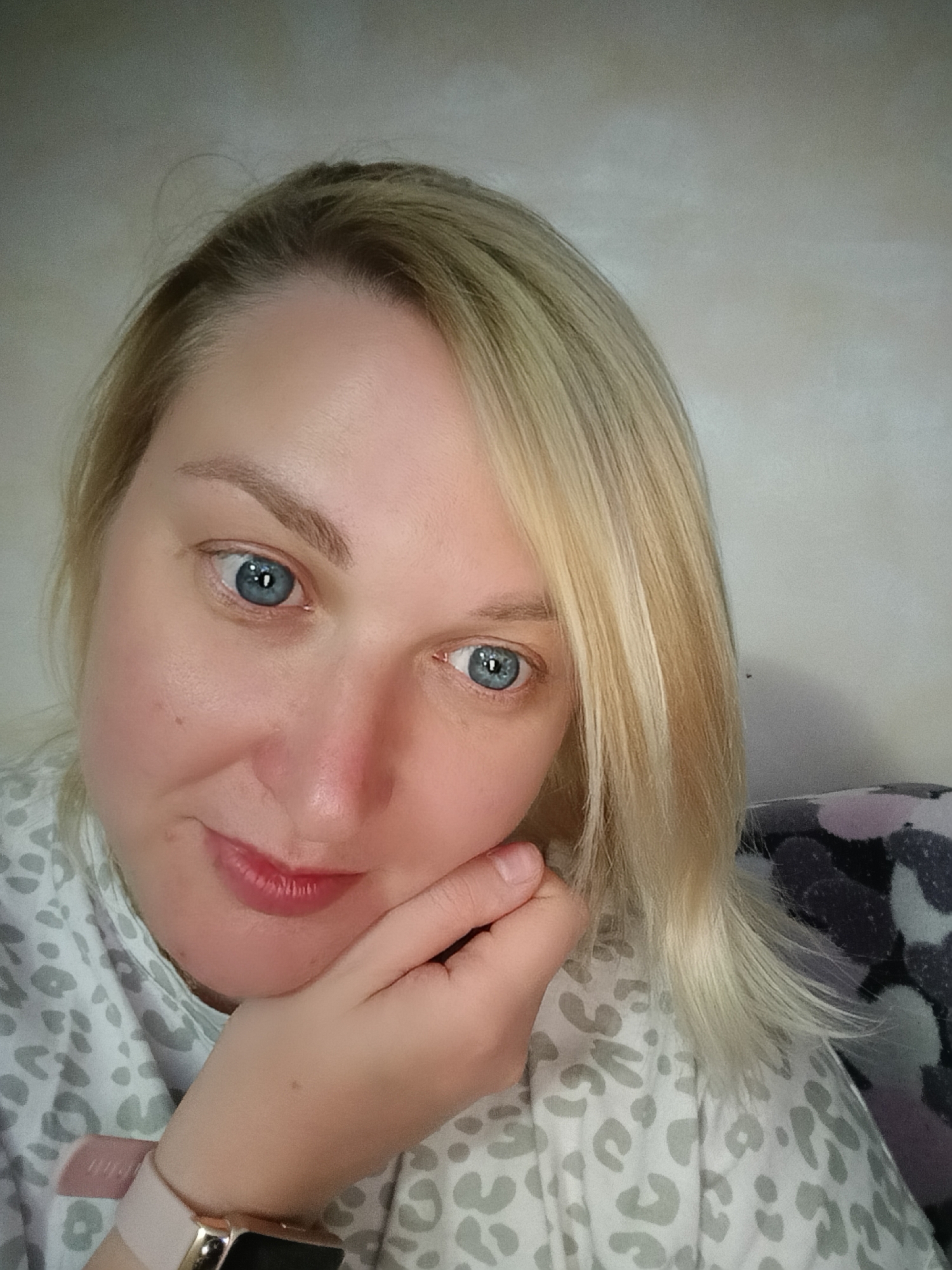 A woman with blonde hair and blue eyes is posing for a close-up selfie. She is wearing a light-colored top with a grey leopard print. Her hand is gently placed against her cheek. A smartwatch with a pink band is visible on her wrist. The background appears to be a plain wall or lightly textured surface. There is a pillow with black, pink, and white print visible to the right of her head. There is no indication of Chaikovsky, Russia, in this photo.