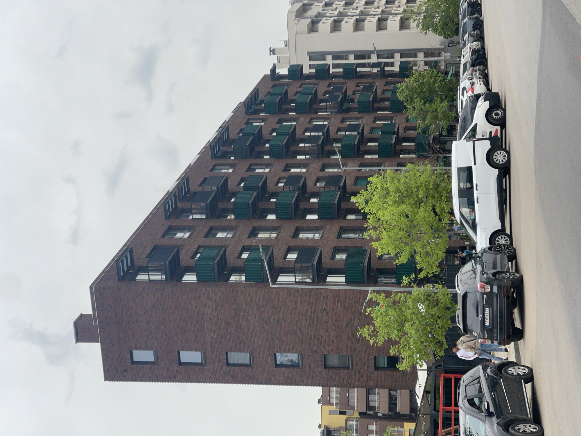 The image shows a multi-story brick building with numerous protruding balconies, likely an apartment complex, in Sofia, Bulgaria. A row of vehicles, mostly vans and SUVs, is parked along the side of the building. A few people are standing near the cars. The sky is overcast.