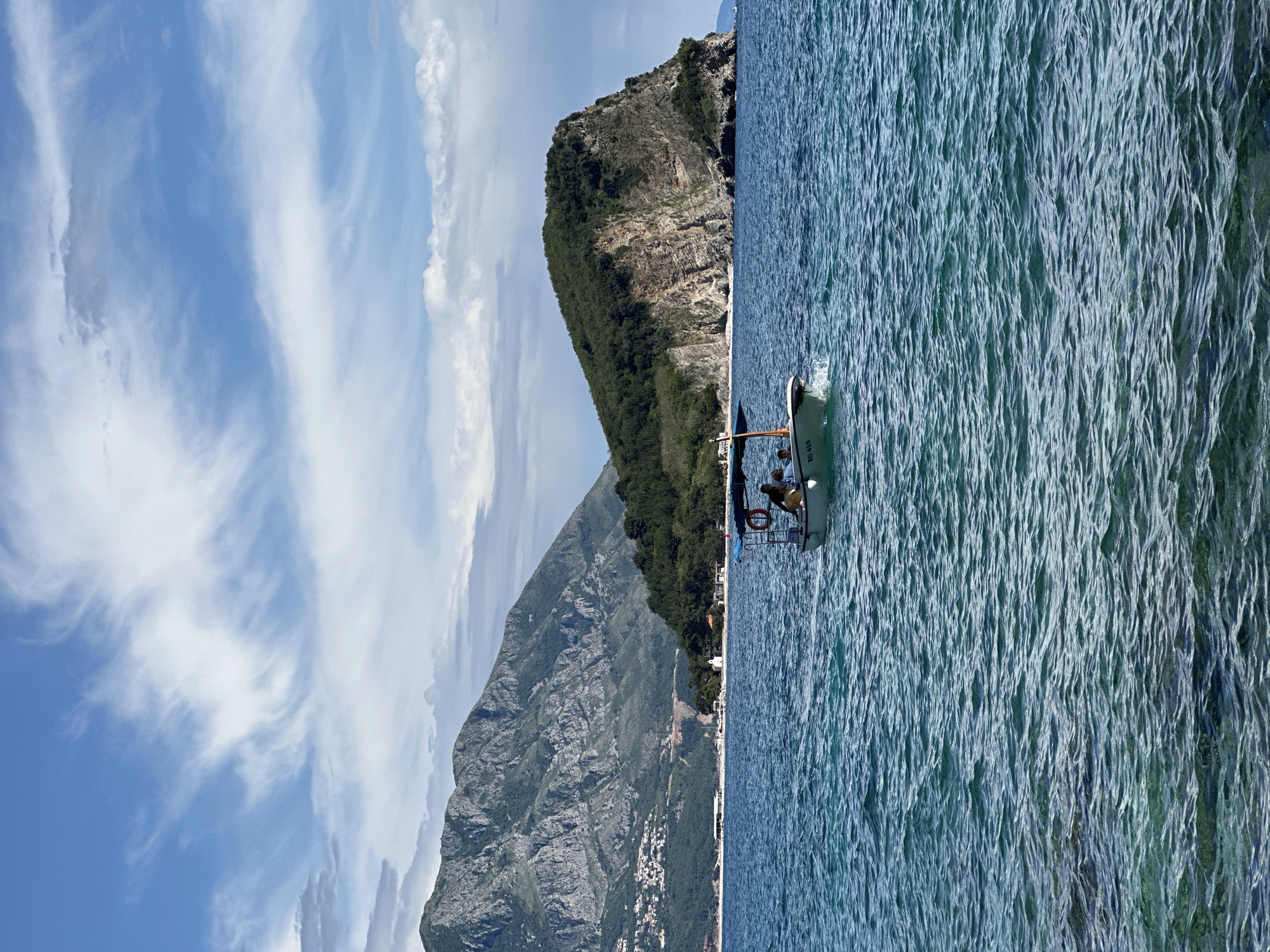 The image captures a scene in Budva, Montenegro, featuring a boat in the sea near the coastline. The sky is blue with scattered white clouds. Mountains covered with greenery rise in the background, with rocky sections visible. A body of water, possibly the Adriatic Sea, fills the right side of the frame, showing its surface texture. A small boat is visible on the water, occupied by what appears to be people, though details are limited due to the distance. The overall scene appears to be taken during daylight hours.