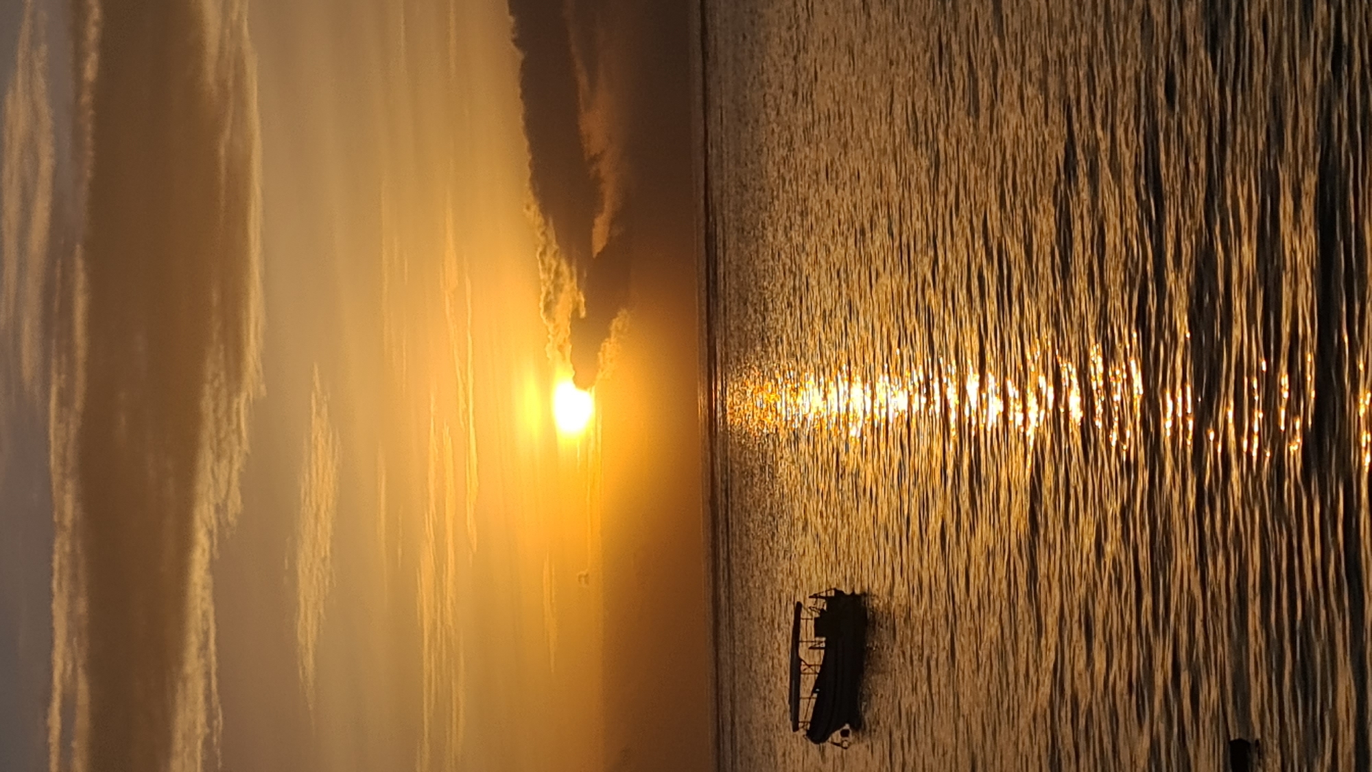 A boat is anchored in calm waters during a sunset over the ocean. The sun is low on the horizon, casting a warm, golden glow across the sky and water. Clouds are scattered across the sky, some tinged with orange and pink from the setting sun. The boat is a small, dark vessel with a canopy. The water is smooth with gentle ripples, reflecting the colors of the sky. The scene evokes a sense of peace and tranquility.