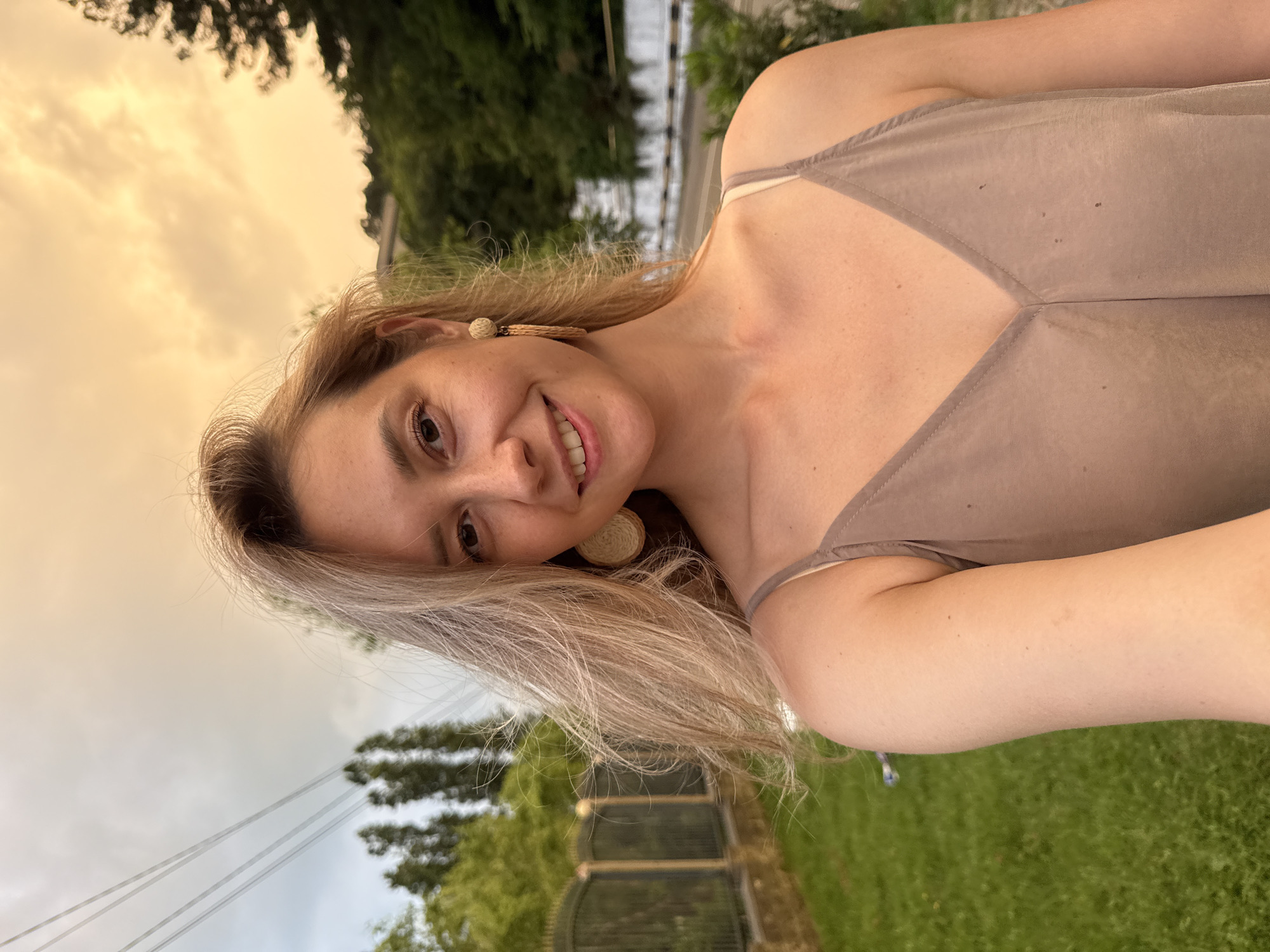 This is a portrait of a young Caucasian woman with long, blonde hair and a warm smile, taken outdoors in the city of Nebug, Russia. The woman is wearing a taupe-colored slip dress and large, circular, woven earrings. She is looking directly at the camera, with her right arm extended to capture the selfie.
The background suggests a suburban or rural setting with elements of nature and infrastructure. To the left of the woman, there is a lush green lawn bordered by a black metal fence. Beyond the fence, tall green trees and some utility poles with wires are visible against a sky with soft, diffused light, possibly indicating dusk or dawn. To the right of the woman, there are more trees and a road with a white barrier, suggesting proximity to a pathway or street. The overall lighting is soft and warm, with the sky displaying hues of orange and yellow.