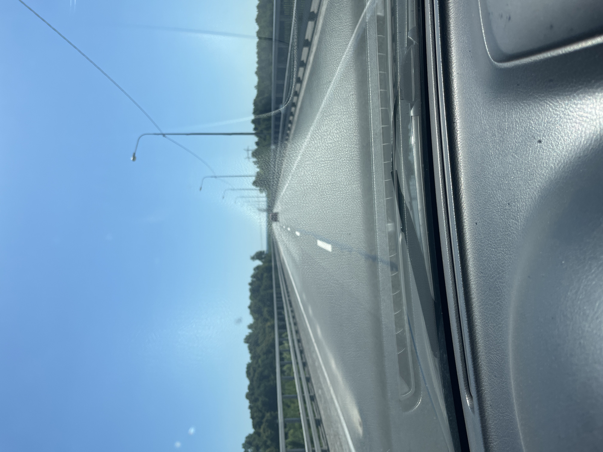 The image shows a view from inside a vehicle, looking out at a highway in Zasur'e, Russia. The perspective is from the passenger side, with a significant portion of the dashboard or interior trim visible on the right. The highway stretches into the distance, with white lane markings clearly visible on the asphalt. On the left side of the highway, there is a line of trees, indicating a rural or semi-rural setting. In the sky, several lampposts can be seen along the side of the road. The sky is a clear, bright blue, suggesting it is daytime, likely mid-morning or afternoon due to the quality of light. There are no people visible in the image. The overall scene depicts travel on an open road under clear skies. The texture of the vehicle's interior is detailed, showing the grain of the plastic material. The image is oriented vertically, with the highway running from the bottom to the top of the frame.