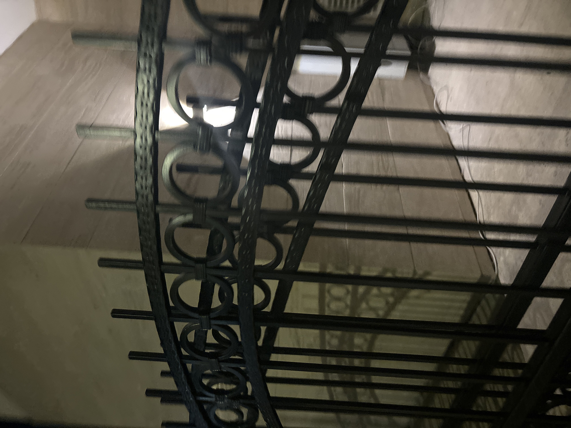 This media file shows a close-up view of a wrought-iron railing, likely part of a staircase or balcony. The railing is dark and intricately designed with circular motifs and horizontal bars. The background appears to be a wall with a tiled or textured surface, possibly in a light beige or tan color. The lighting suggests it might be indoors or at night, with a bright light source creating a strong highlight on the railing.
The perspective is angled, looking up towards the railing. The image is slightly out of focus, giving it a soft, almost dreamy quality. There are no people visible in the frame. The scene depicts an architectural detail, focusing on the craftsmanship of the metalwork. The repetition of the circular elements and the linear structure of the horizontal bars create a sense of pattern and rhythm.
The context provided is Budva, Montenegro, which might imply the railing is part of a historic building, a hotel, or a residential property in that location. However, there are no explicit visual cues in the image itself to confirm this specific location, such as signage or unique architectural styles beyond the railing. The overall mood is neutral, with the focus being on the visual texture and design of the railing. No text is visible in the image.