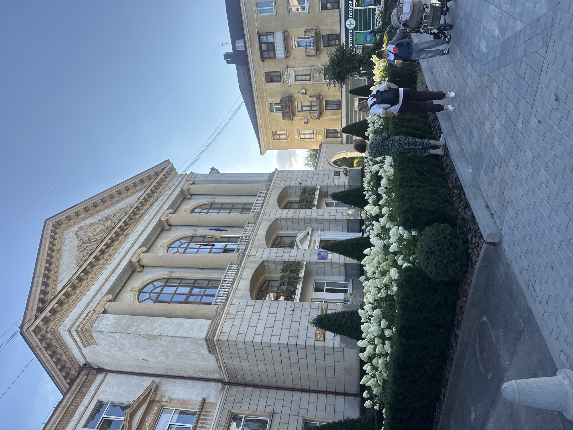 The image shows the Ternopil Regional State Administration building, a grand, classical-style structure with a light-colored stone facade, large arched windows, and ornate architectural details. In front of the building, there is a well-maintained garden with manicured hedges and a profusion of white hydrangeas. A paved walkway runs along the garden. Three individuals are visible on the walkway. On the left, a woman in a dark floral dress stands facing away from the camera. In the center, another woman wearing a white t-shirt and dark pants with a backpack stands with her back to the camera. To the right, a young boy in a grey jacket and blue jeans holds a yellow cup and stands next to a stroller. The sky is clear and blue, with some thin power lines visible. To the right of the administrative building, a yellow apartment building with multiple balconies is visible, and a green sign with a pharmacy logo and text in Ukrainian can be seen on its ground floor.