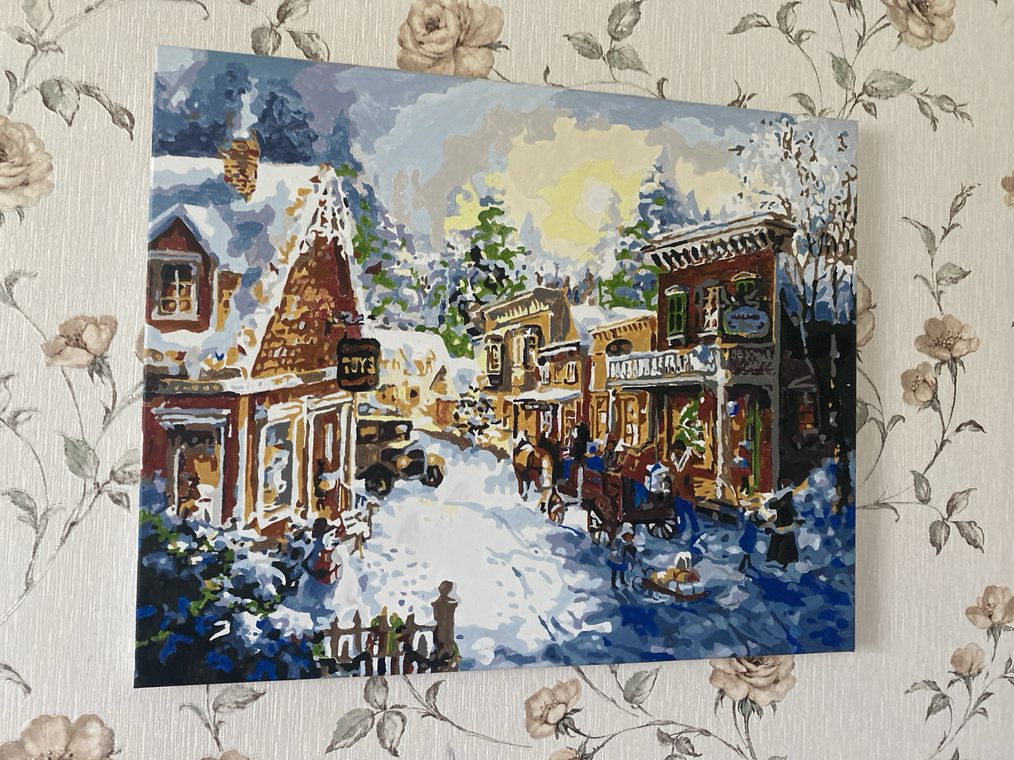 The image displays a painting of a snow-covered town street with buildings on either side. The style of the painting appears to be a paint-by-numbers artwork. The left side of the street features a building with a red roof and a sign that reads "TOYS". There is a horse-drawn carriage with people in it on the street. Further down the street, more buildings are visible, with a bright, yellowish sky in the background. The painting is hanging on a wall with floral wallpaper. The image does not contain any information about the city of Vvedenka, Ukraine.