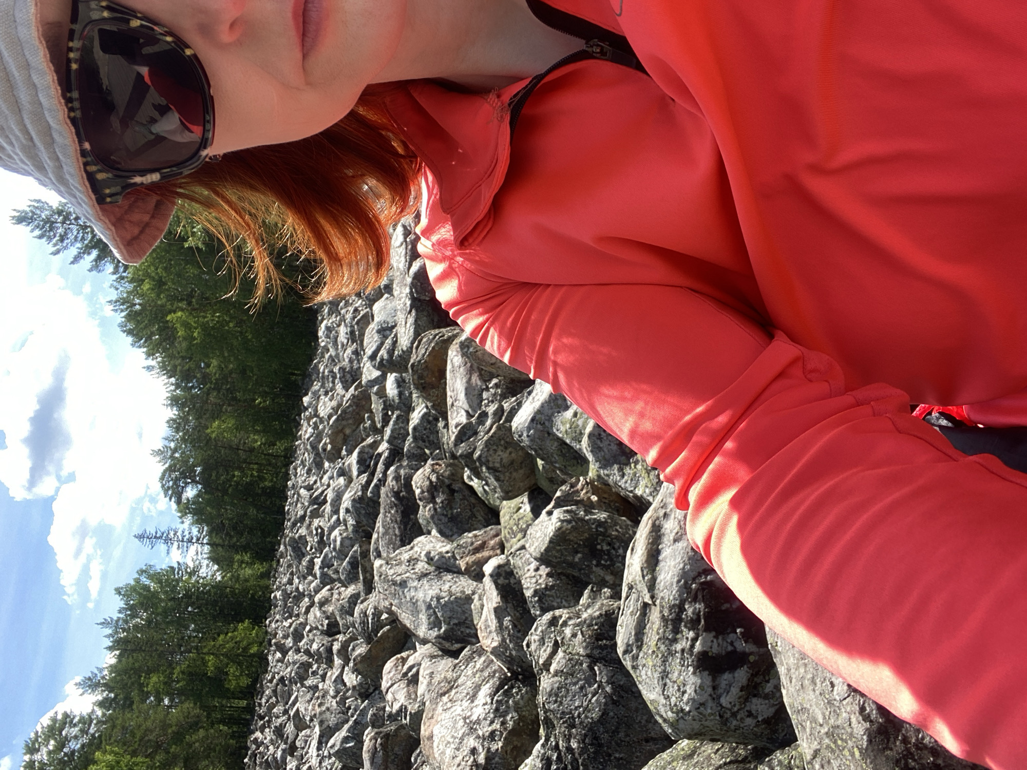 A person with red hair, wearing sunglasses, a bucket hat, and a bright red jacket, takes a selfie in a rocky outdoor area. The background shows a dense forest and a partly cloudy sky. The ground is covered with numerous grey rocks of varying sizes, interspersed with some vegetation. The person's face is partially visible, and their arm and torso are in the foreground. The image appears to be taken on a sunny day, likely during the summer. The location is identified as the Taganay National Park, within the city of Zlatoust, Russia.