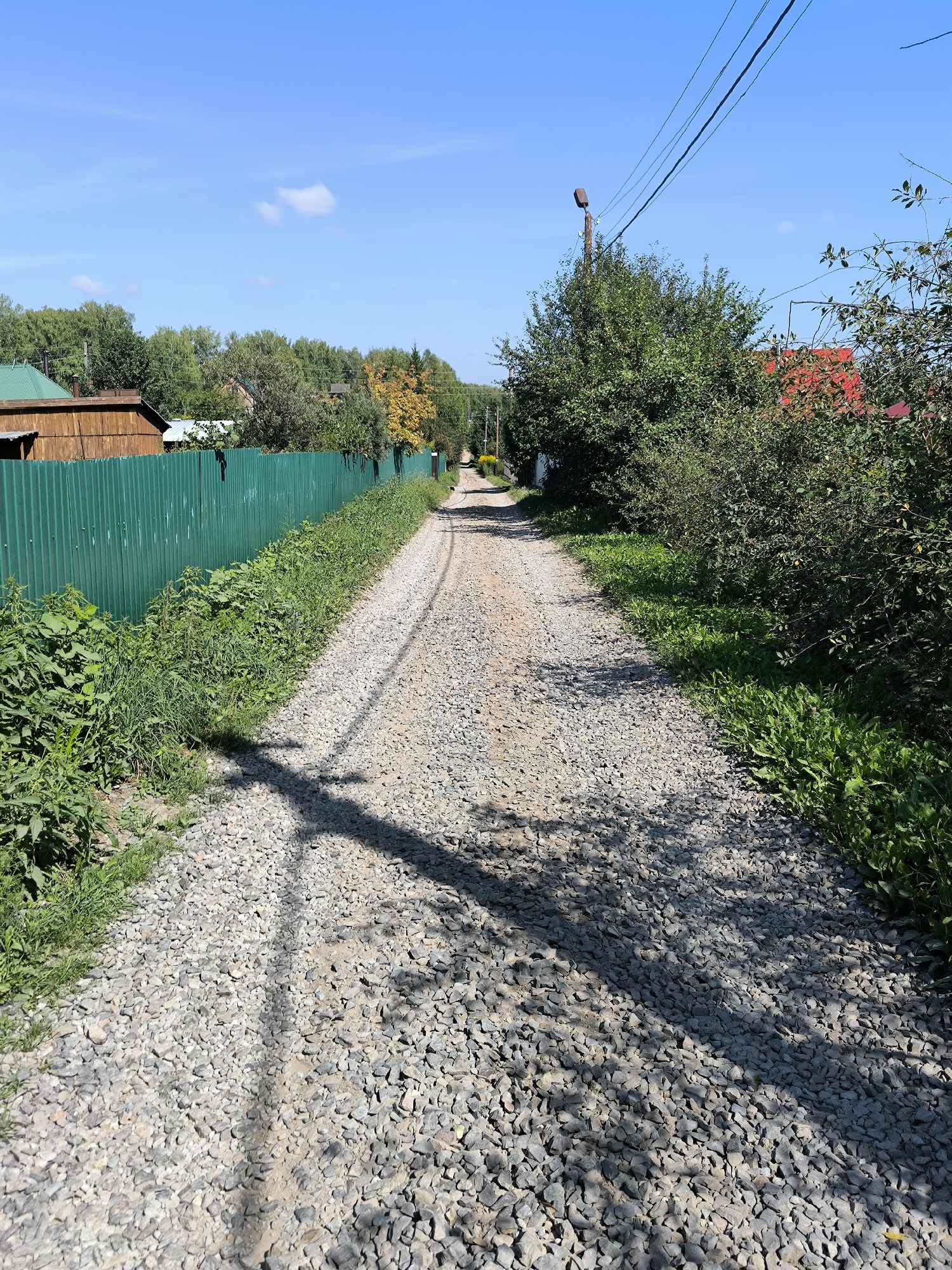 The image depicts a gravel road winding through a rural area, likely a dacha settlement, in Maiskii, Russia. The road is flanked by a tall, green corrugated metal fence on the left, beyond which are wooden buildings with green roofs. Lush green vegetation grows along the fence and the roadside. On the right, a dense growth of trees and bushes lines the road, with glimpses of red-tiled roofs and more trees further in the background. Power lines stretch across the clear blue sky, supported by a wooden pole with a light fixture. The sunlight casts distinct shadows from the trees onto the gravel road, creating a dappled effect. The scene suggests a quiet, sun-drenched day in a semi-rural environment.